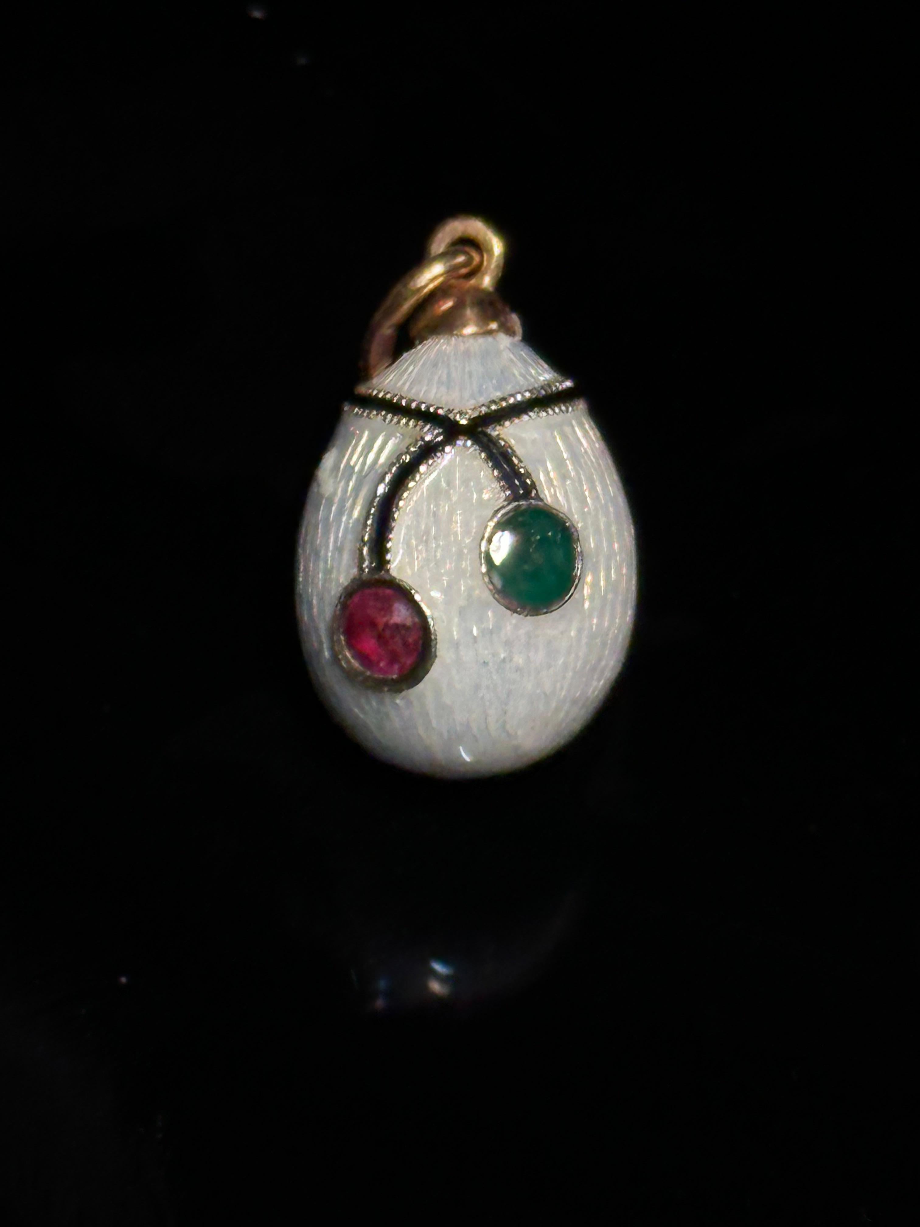 Antiguo colgante de huevo de esmalte blanco al estilo Fabergé, con esmalte blanco y detalles de rubíes y esmeraldas. La base es de plata de ley 585, mientras que los detalles dorados de ley 750 añaden una elegancia especial. El peso total del