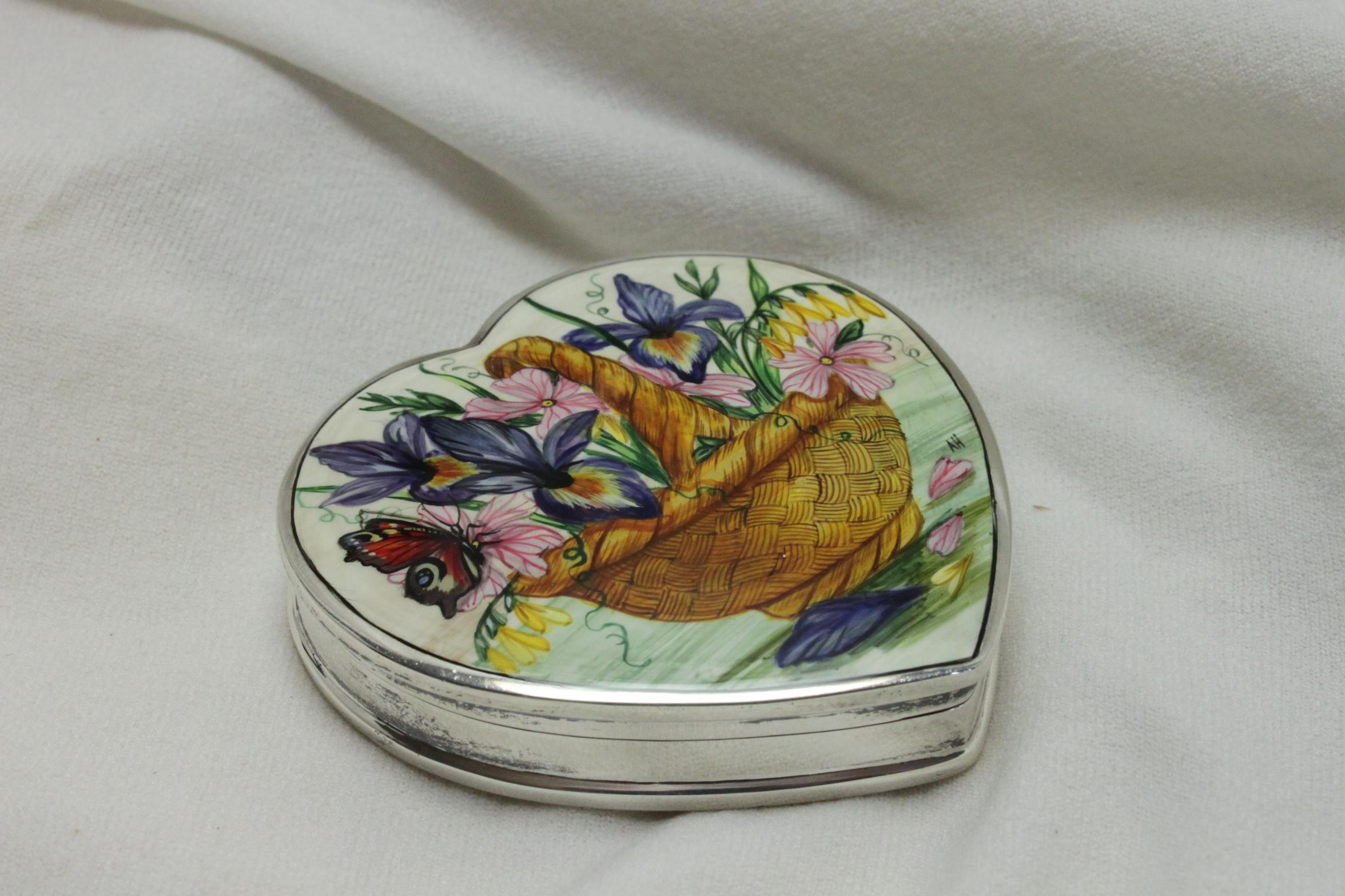 Dieses hübsche herzförmige Schmuckstück aus Sterlingsilber ist mit einer handgemalten Szene eines Weidenkorbs voller Blumen und einem Schmetterling, der auf einer der Blüten ruht, verziert. Das Gemälde ist mit AH signiert. Es misst 66 mm in der