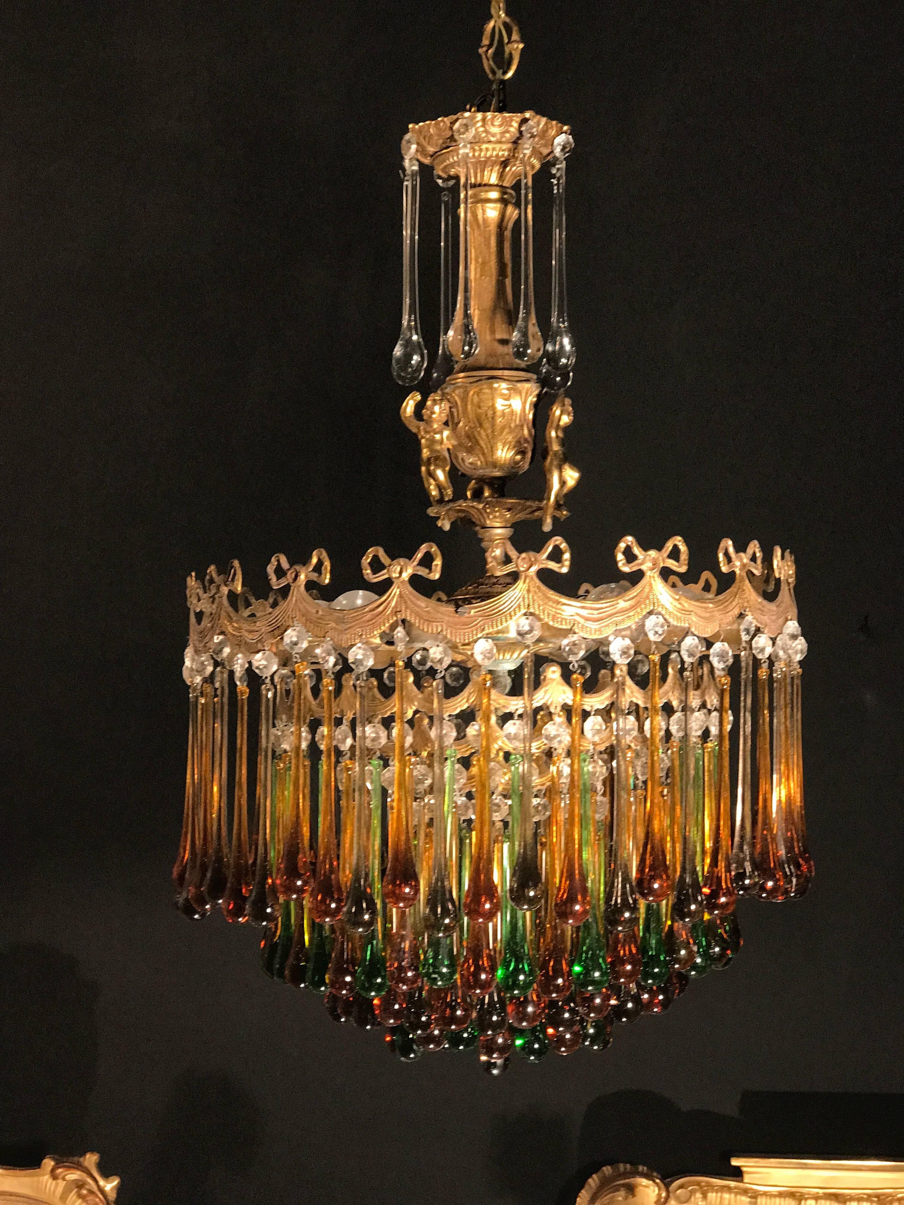 Splendid waterfall chandelier with a finely chiseled brass frame with four lower tiers all of which are dripping a multicolored teardrop glasses. The center is adorned with three graceful dancing putti.
There are six E 27 light bulbs.
Supplied