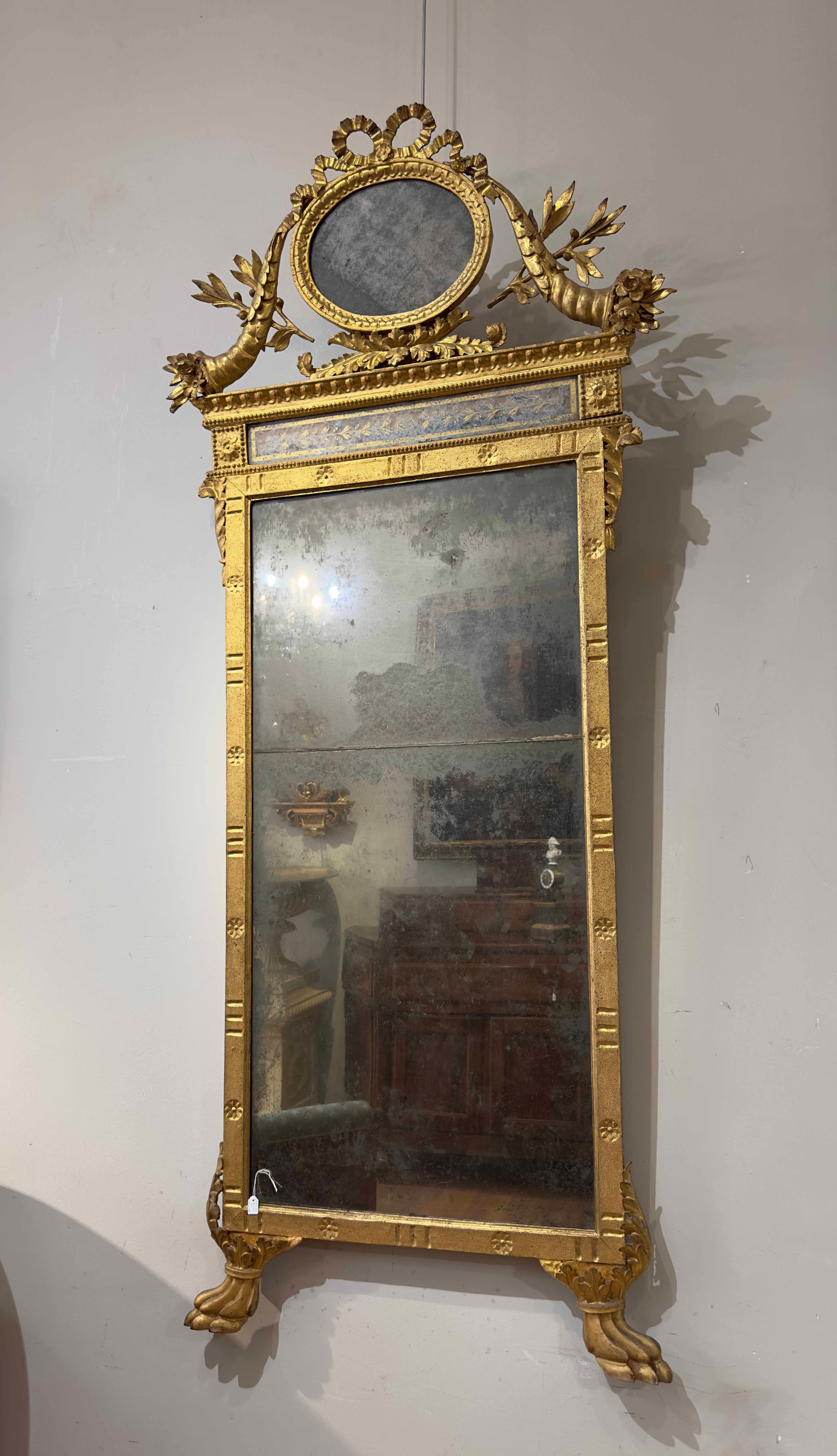Beautiful and elegant neoclassical mirror in carved and gilded pine wood with pure gold leaf. The cymatium features decorations that recall abundance and peace, with cornucopias and olive branches. The upper termination of the cymatium is instead