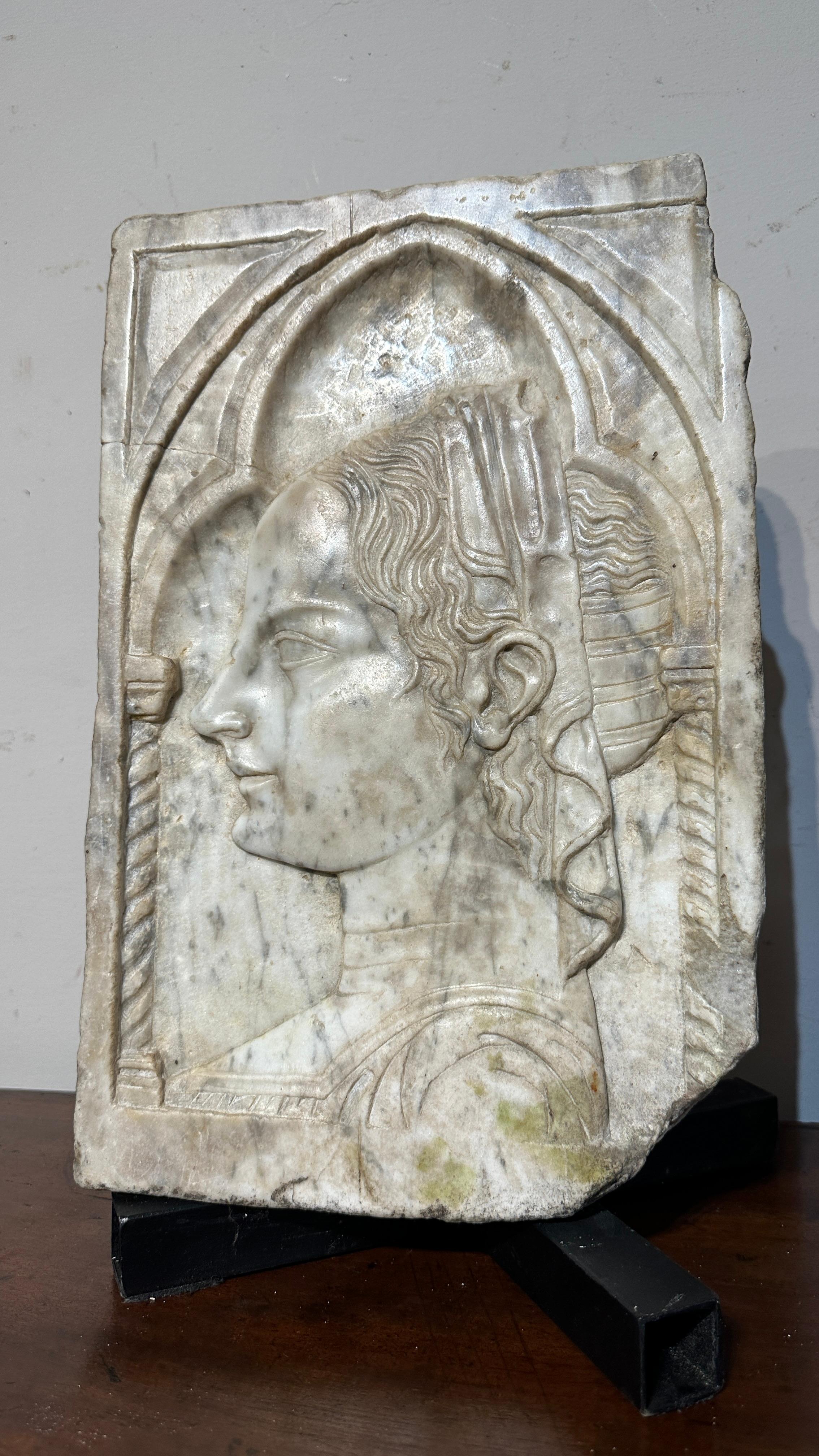 Beautiful carved white marble plaque, depicting a noblewoman with elegant hair typical of the 14th century and inside a trefoil arch, supported by twisted columns. The plaque can be attributed to Alceo Dossena, a famous artist active between the end