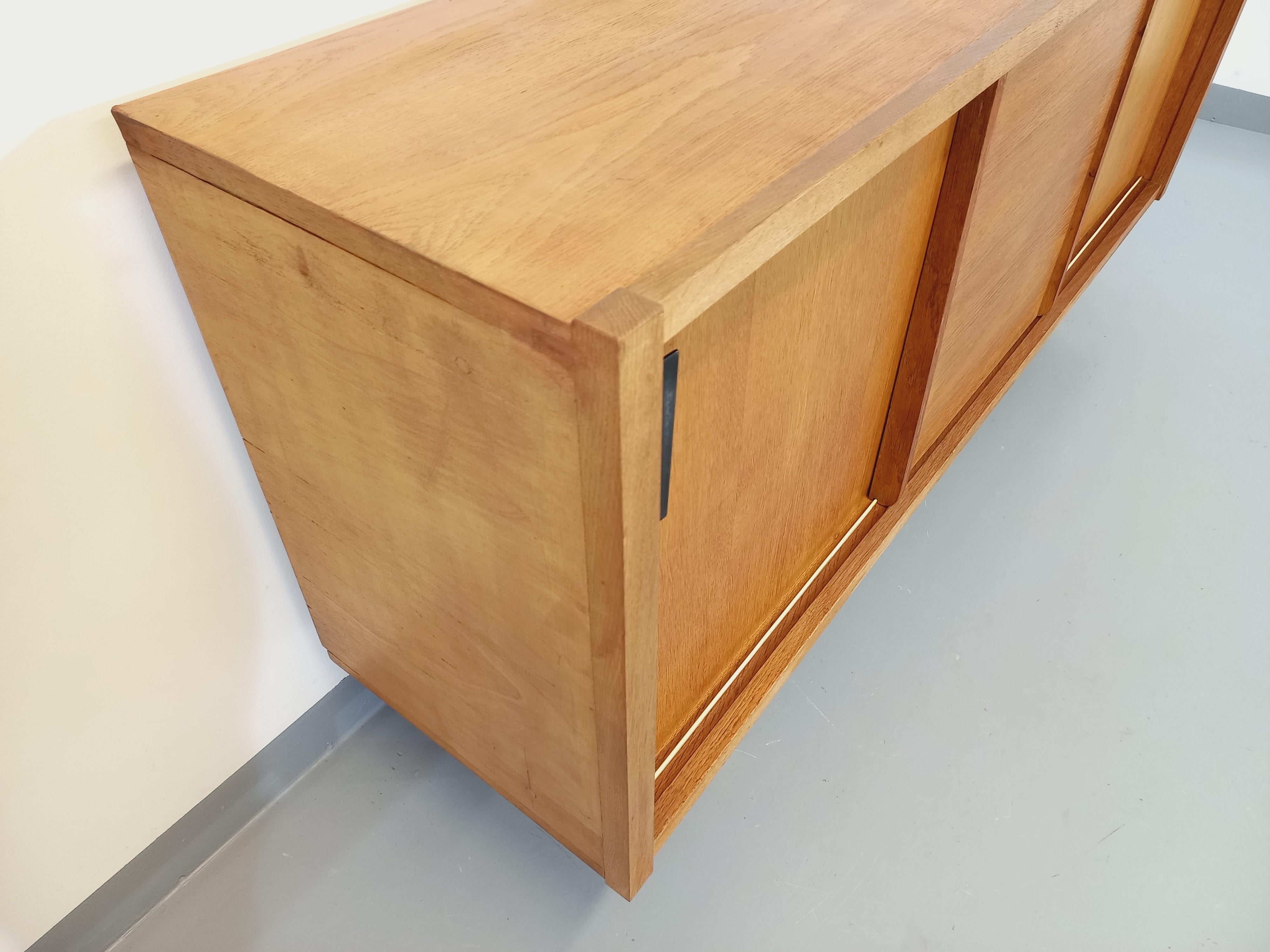 Enfilade vintage des années 60, en bois de chêne blond doré, très beau design moderniste avec poignées asymétriques en métal noir et pieds compas. Beaucoup de rangements : 3 portes coulissantes avec diverses étagères et casiers intérieurs. Très bon