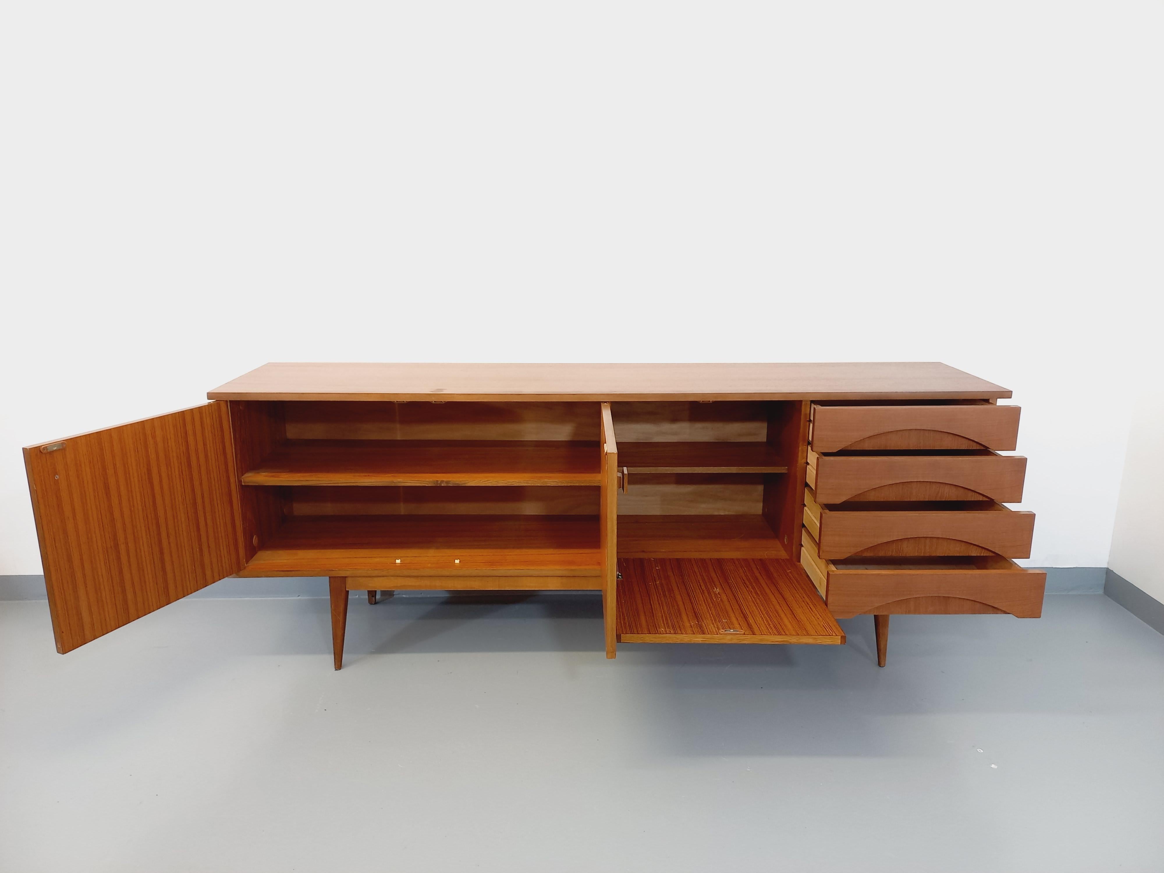 Enfilade basse vintage style scandinave des années 60, en teck, pieds compas, jolies poignées carrées. Beaucoup de rangements : 2 portes avec étagère, 1 partie bar avec porte abattante et étagère, 4 tiroirs avec de belles découpes. Très bon état