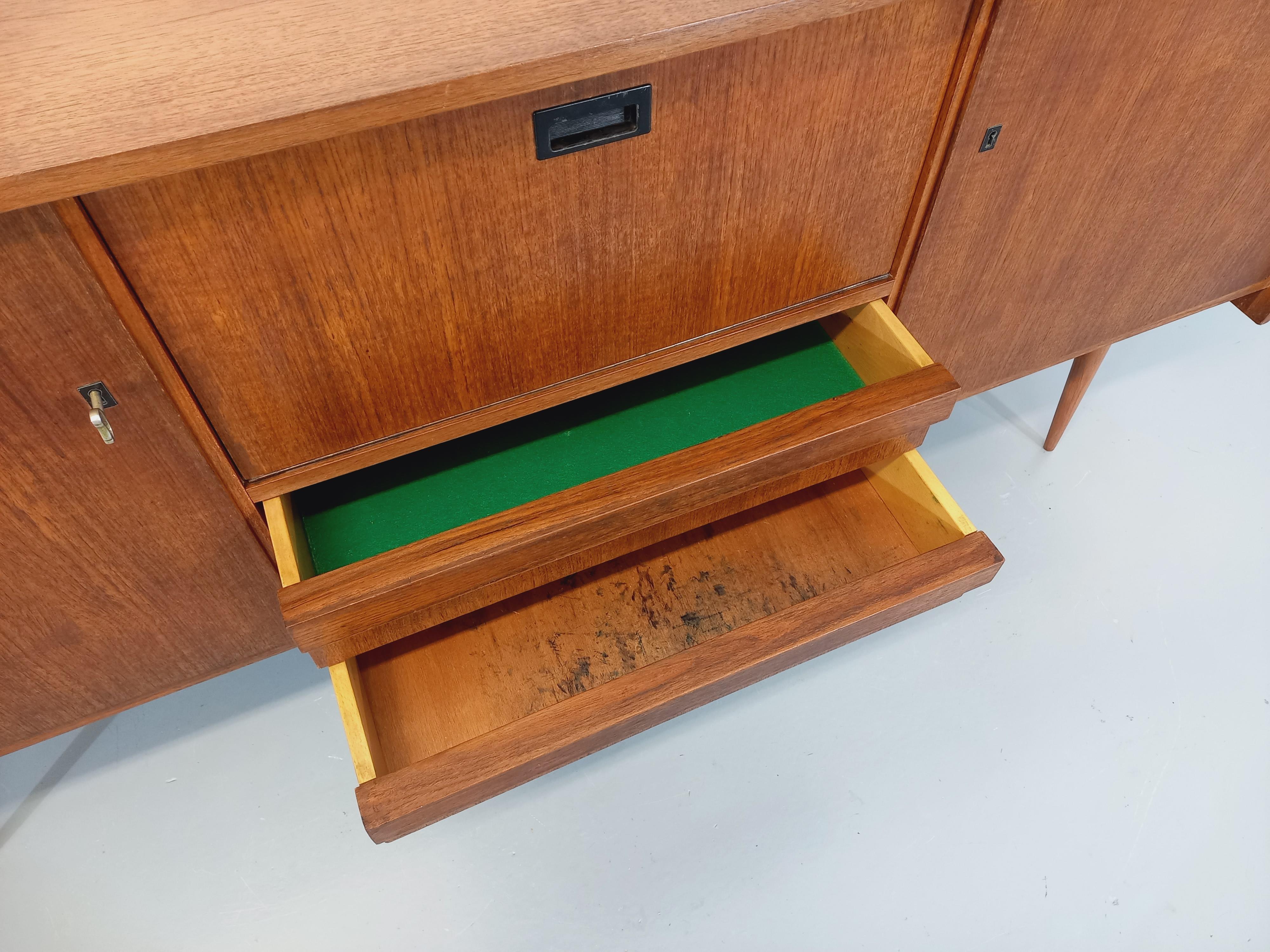 Enfilade vintage style scandinave des années 60, en teck, très beau design aérien avec pieds compas en hauteur. Beaucoup de rangements : 2 portes avec étagères et clé, 2 tiroirs, 1 partie bar avec porte abattante à poignée noire et miroir en fond.