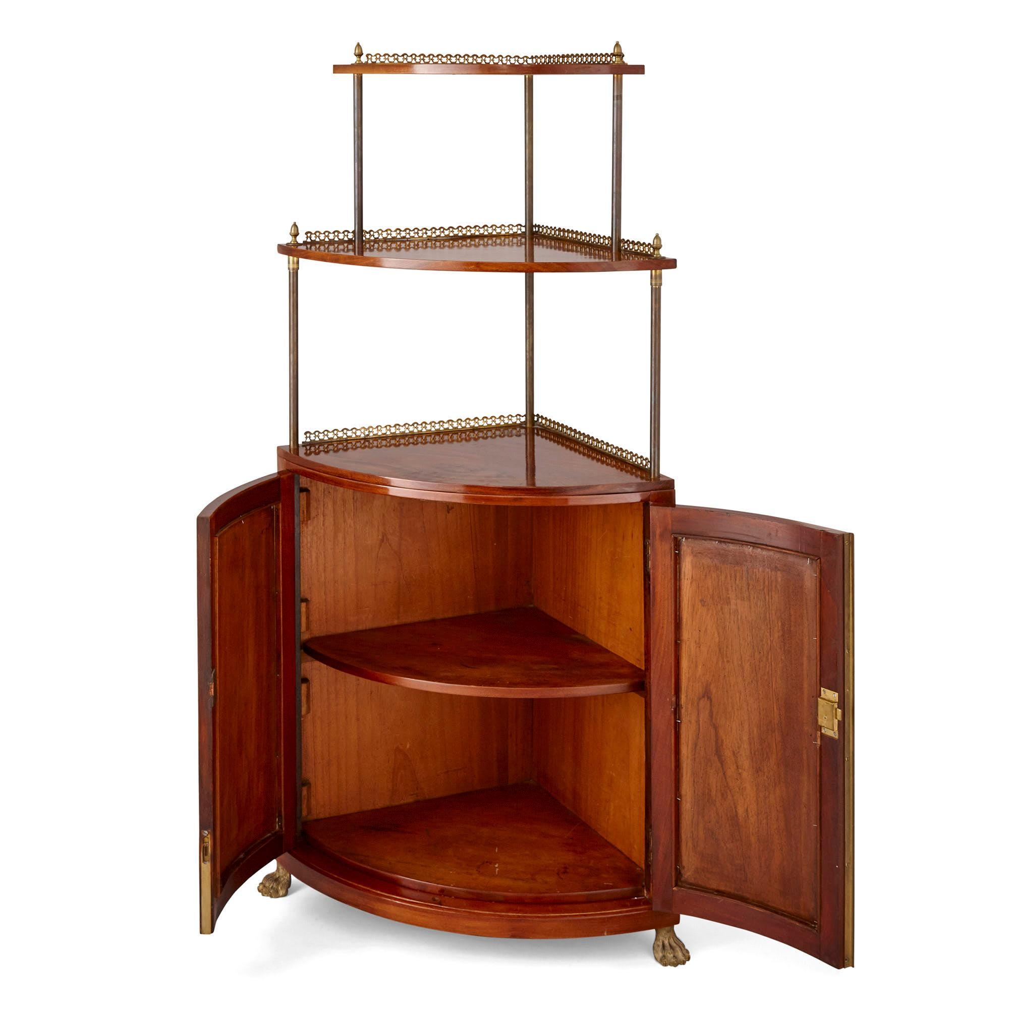 This corner unit, crafted from mahogany and gilt bronze, functions as a cabinet and a tiered shelf. The cabinet, quarter-circle shaped in profile, is supported by paw feet and features, to the front, twin curvilinear doors. The doors, which open to