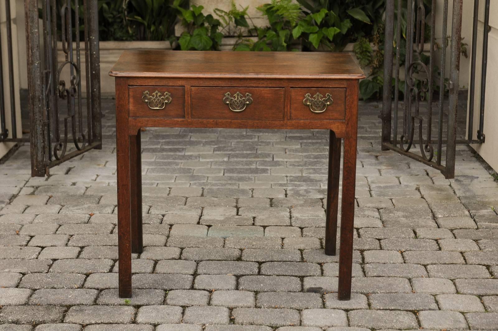 An English Georgian period oak side table from the late 18th century, with three drawers, chinoiserie hardware and Marlborough legs. This English oak side table features a rectangular planked top with molded edges and rounded corners in the front,