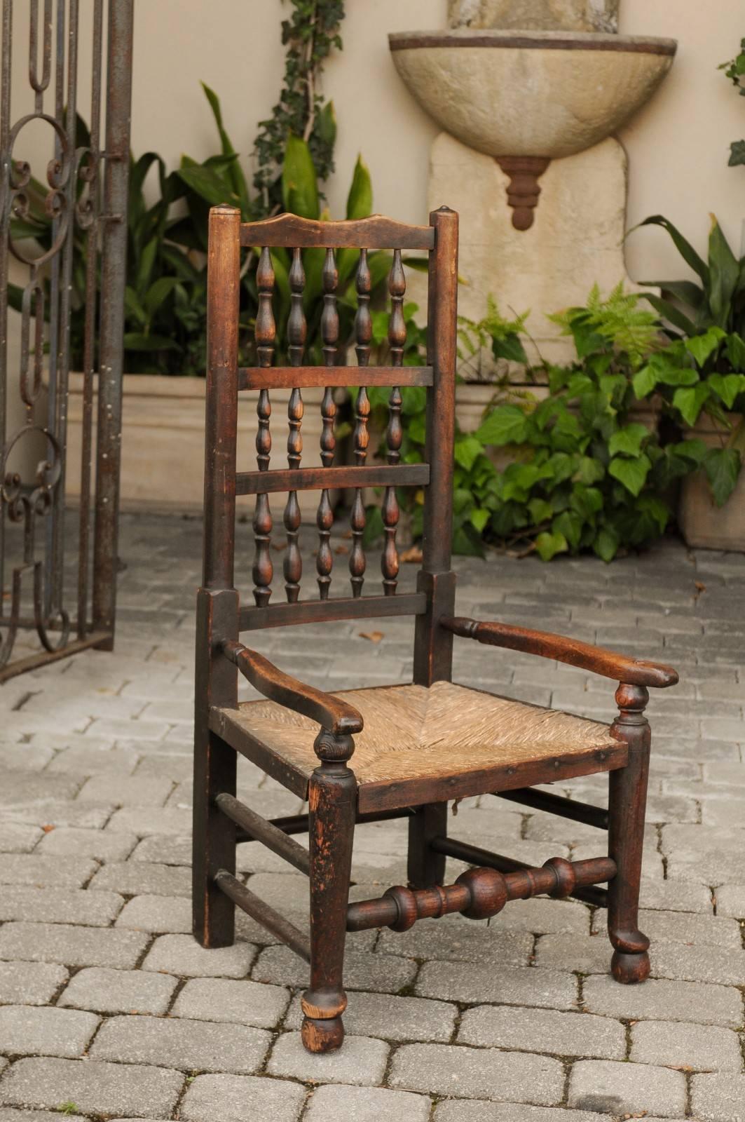 An English William and Mary style oak youth chair from the late 18th century, with rush seat and spindle back. This English oak chair features a straight spindle back, connected to two open arms, adorned with delicately splayed extremities. The