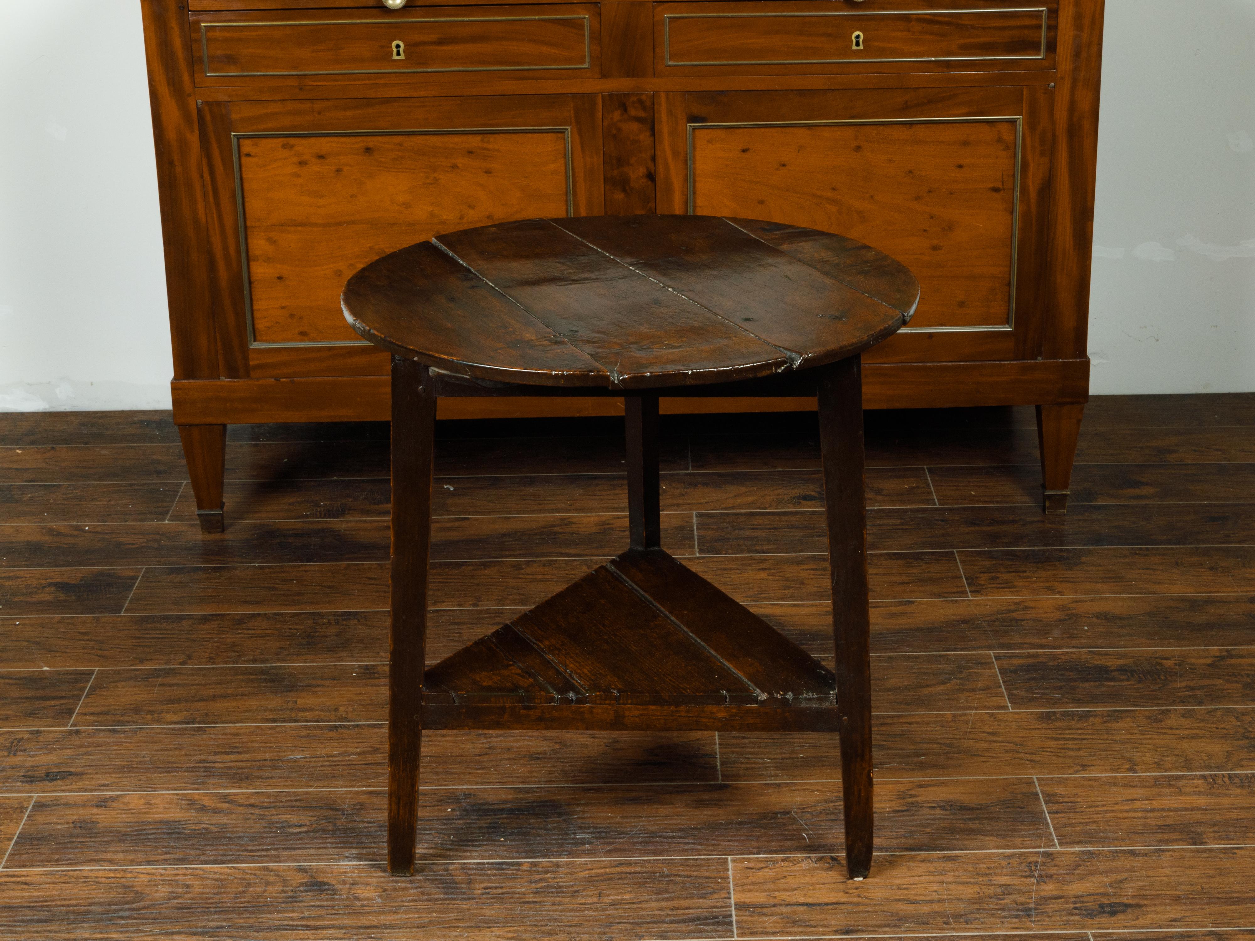 An English oak cricket table from the early 19th century, with circular top, triangular shelf and dark patina. Crafted in England during the early years of the 19th century, this rustic cricket side table features a round top sitting above a simple