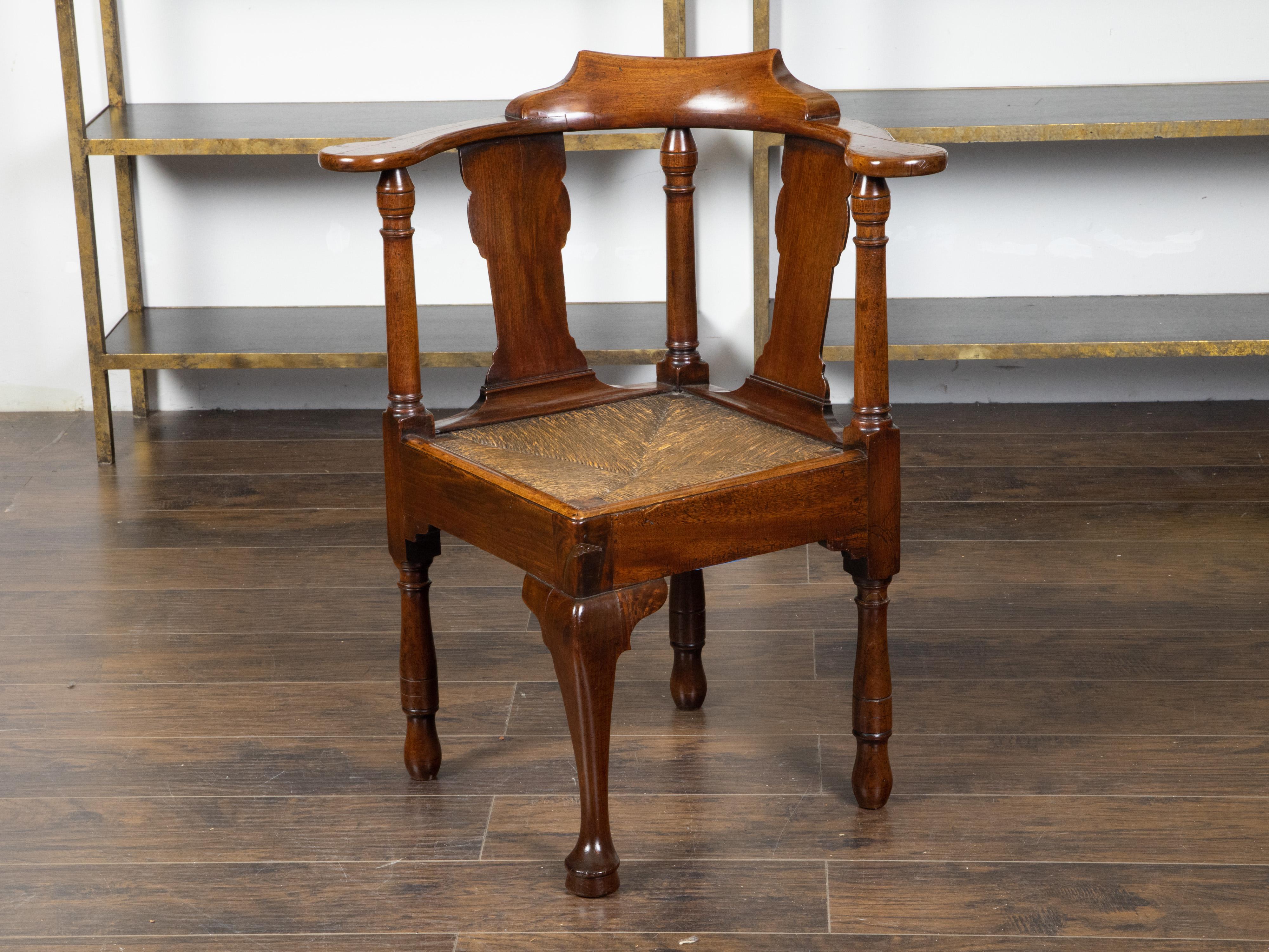 An English mahogany corner chair from the early 19th century, with carved crest, rush seat and front cabriole leg. Created in England during the first quarter of the 19th century, this mahogany corner chair features a pierced back alternating carved