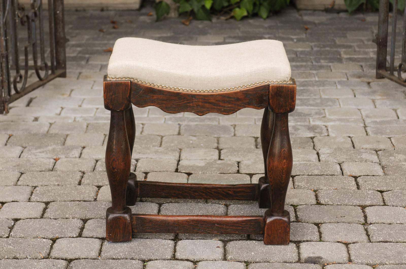 An English oak saddle seat stool from the early 19th century with baluster legs, side stretchers, new upholstery and brass nailheads. This English oak stool features an exquisite saddle seat, covered with a new linen upholstery, accented with a