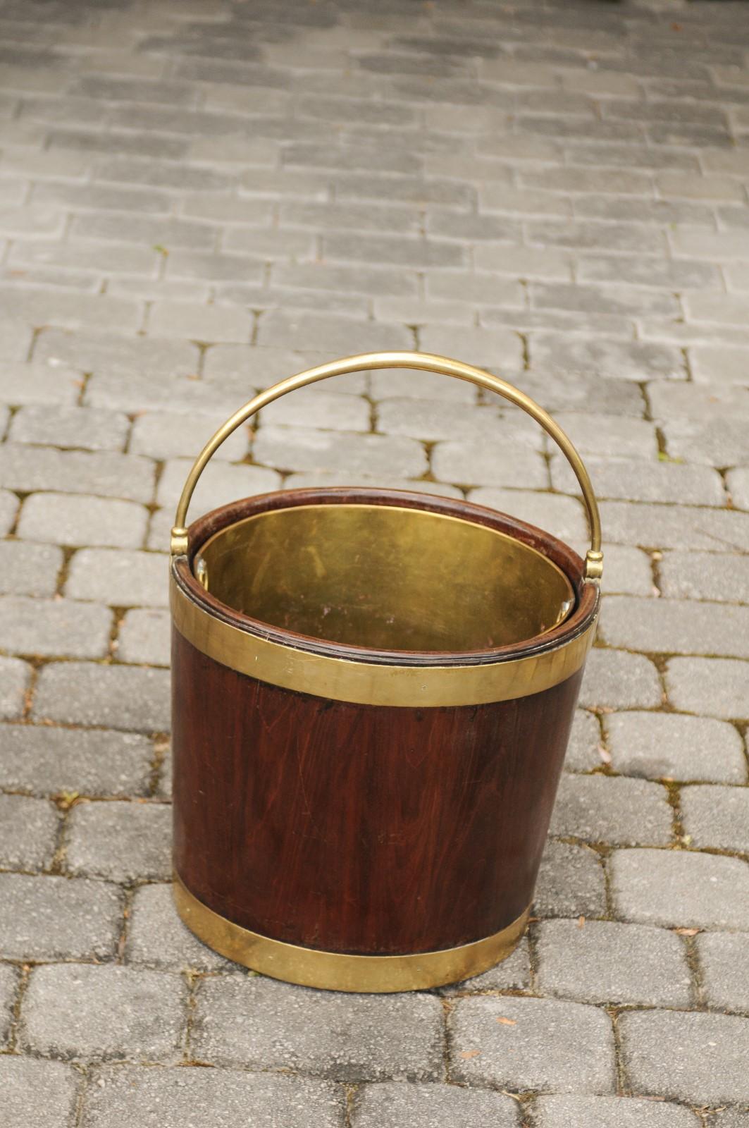 An English Regency mahogany bucket from the early 19th century, with brass accents and handle. Born in England during the second quarter of the 19th century, this elegant mahogany bucket features a removable brass liner. Topped with a large handle