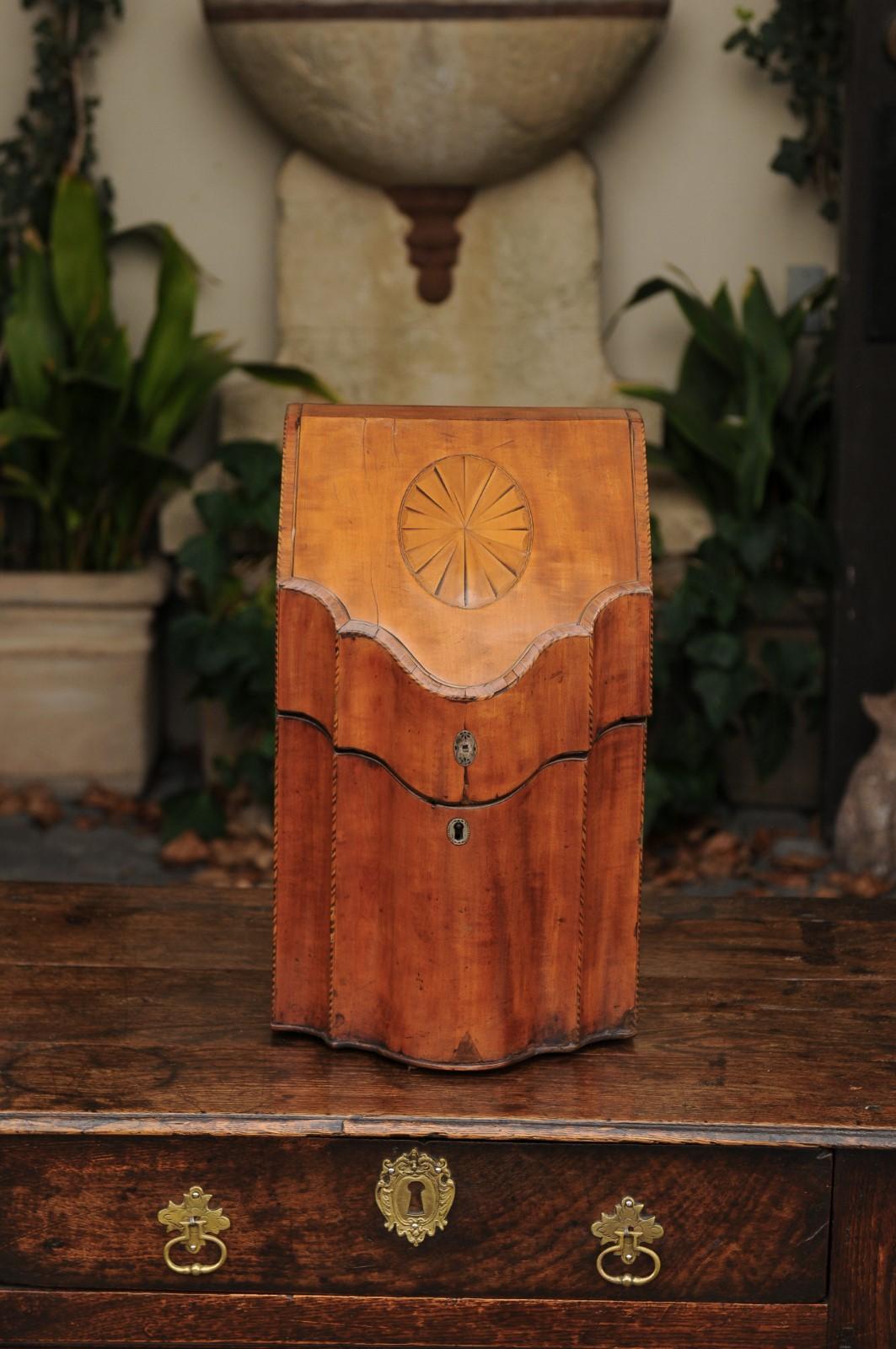 An English mahogany knife box from the first half of the 19th century, with serpentine front, checkered stringing and inlay. This exquisite English knife box presents a brass hinged top adorned with an oval inlaid medallion opening to reveal an