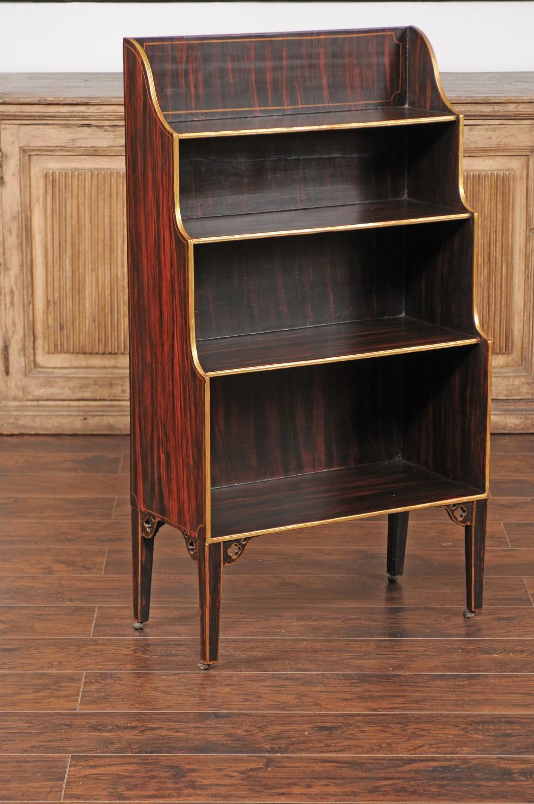 An English faux-painted waterfall bookcase from the mid-19th century, with gilded accents and tapered legs. This English faux-painted waterfall bookcase from the mid-19th century exudes a charming blend of functionality and decorative flair. Crafted