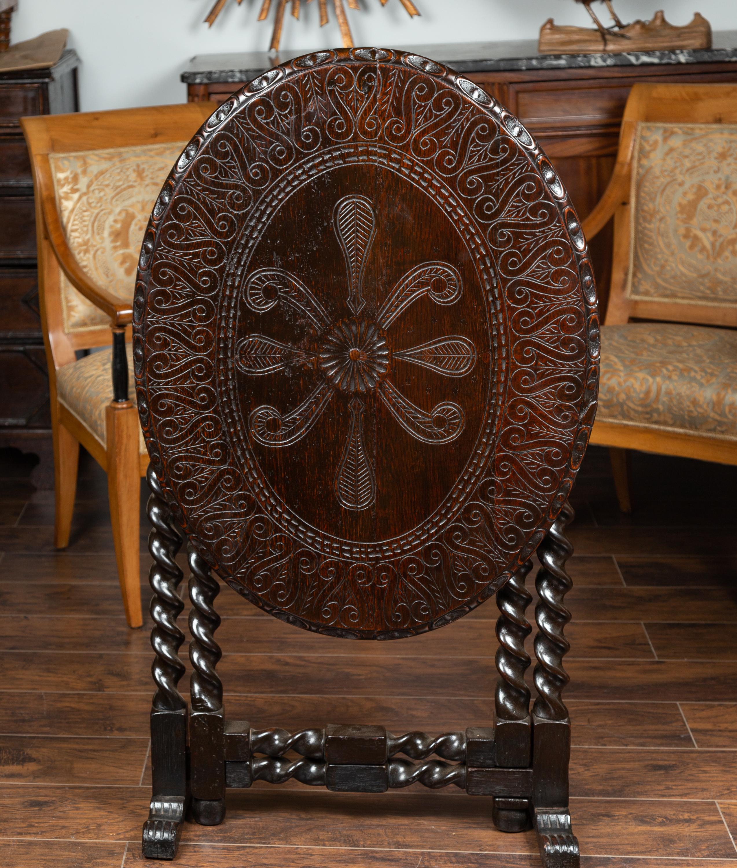 An English oak gate leg oval table from the mid-19th century, with carved top and barley-twist base. Born in England during the mid-19th century, this table captures our attention with its oval tilt-top, carved with delicate volutes and foliage