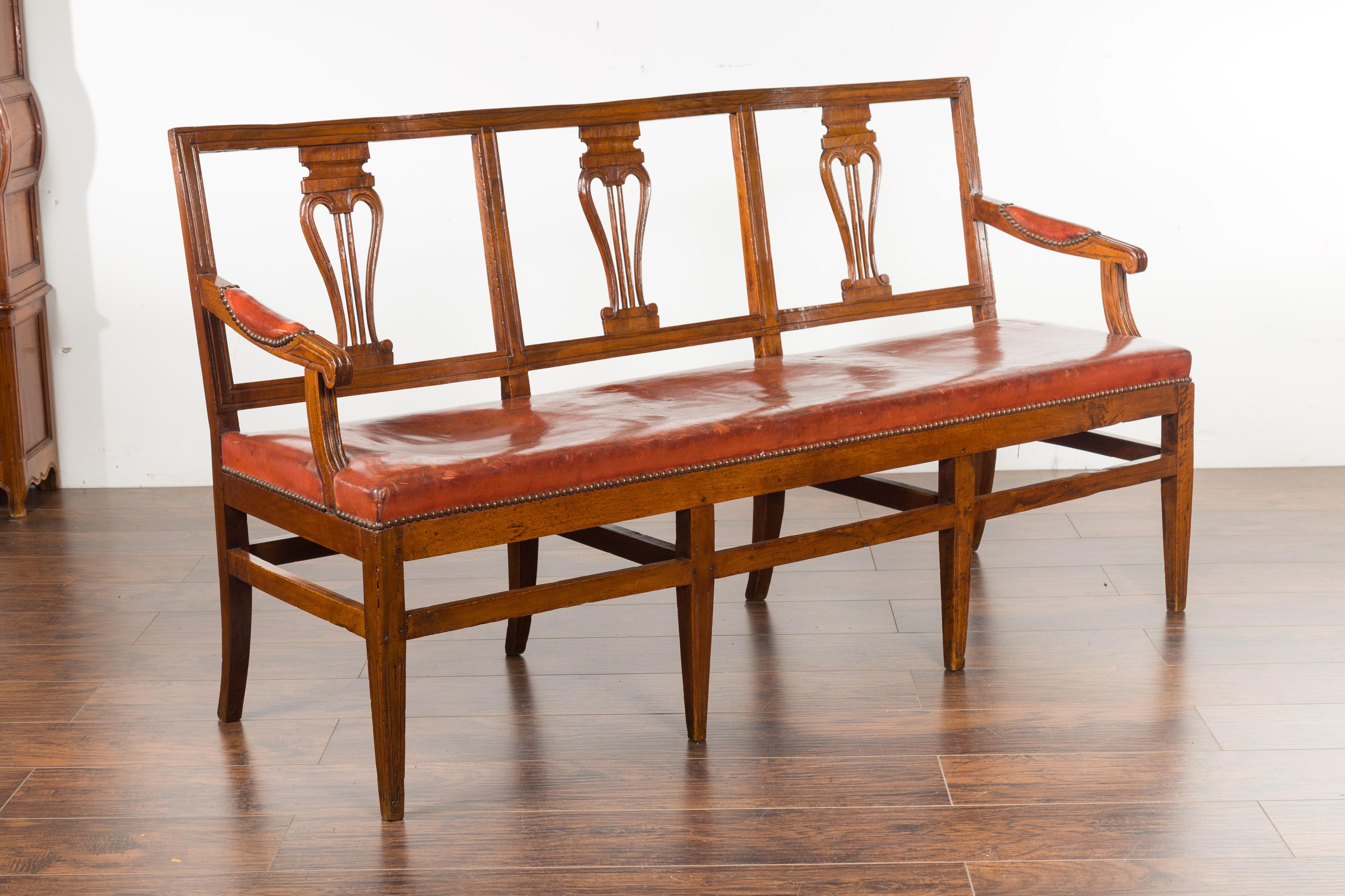 An English walnut three-seat bench from the mid-19th century, with leather seat and carved back. Created in England during the third quarter of the 19th century, this walnut bench features a pierced back adorned with delicate volutes on the splats,
