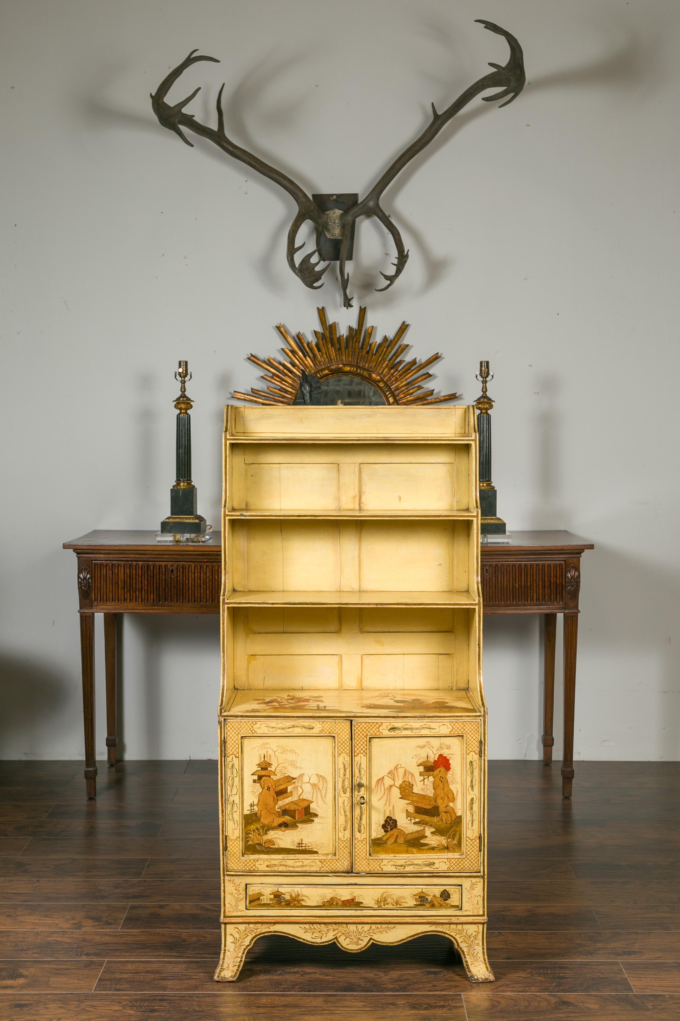 An English painted Chinoiserie waterfall bookcase from the late 19th century. Attracting our eyes with its soft yellow painted color adorned with Chinoiserie landscape motifs, this waterfall bookcase features open shelves of increasing sizes sitting