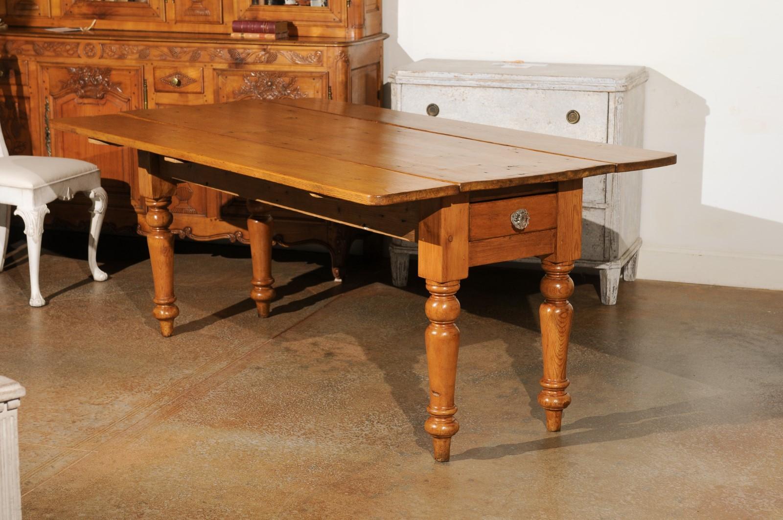 An English pine drop leaf farm table from the late 19th century, with lateral drawers and turned legs. Created in England during the third quarter of 19th century, this farm table features a rectangular top flanked with two drop leaves, measuring
