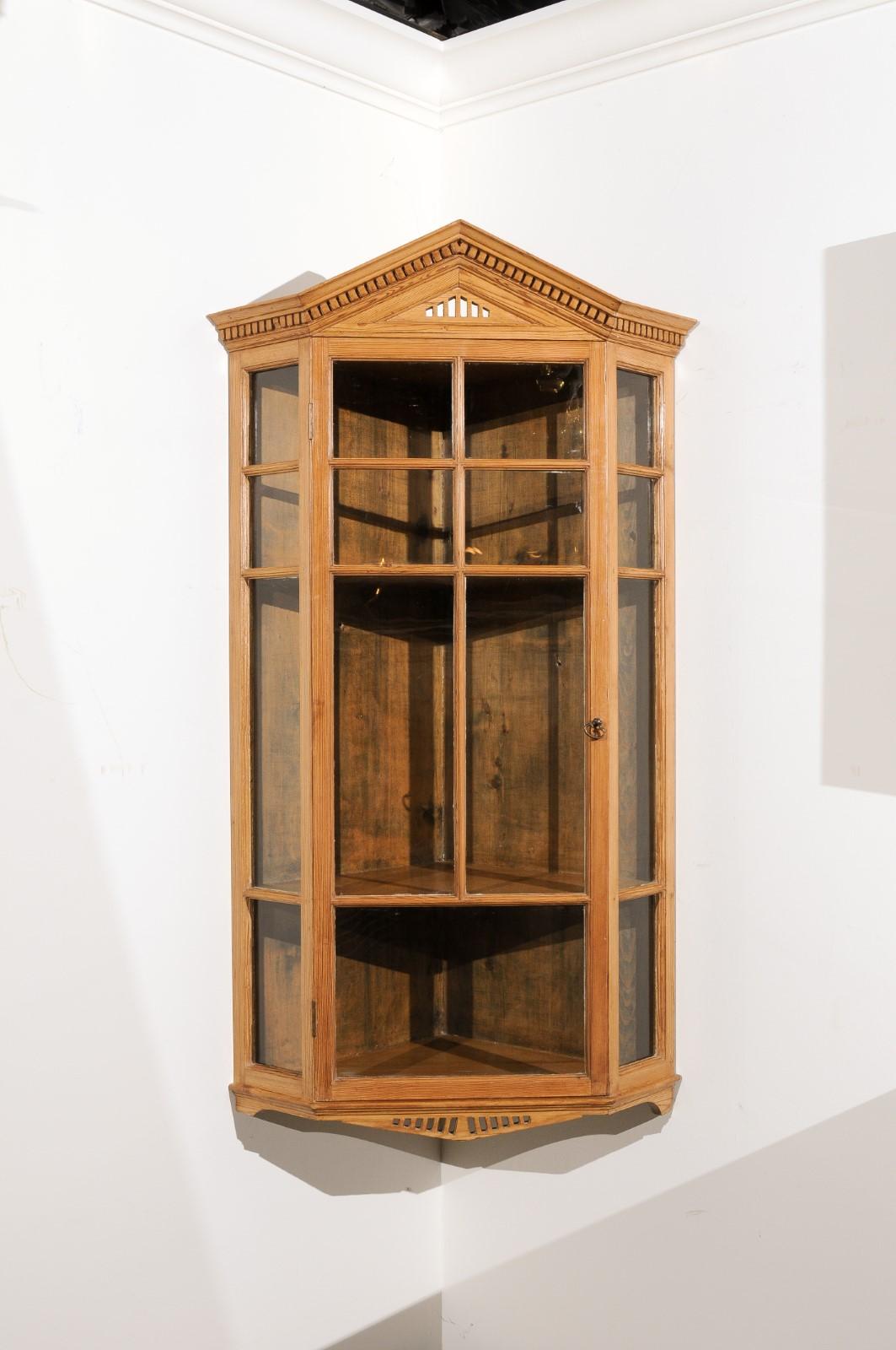 An English pine hanging corner cabinet from the late 19th century, with pointed pediment, dentil molding, glass doors and canted corners. Created in England during the third quarter of the 19th century, this pine wall hanging corner cabinet captures