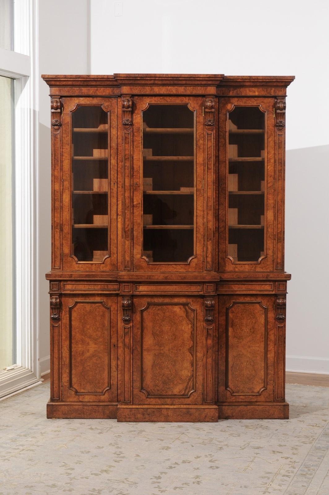 An English burled walnut breakfront bookcase from the late 19th century, with glass doors and carved volutes. Born in England in the last quarter of the 19th century, this splendid bookcase features a burled walnut structure, topped with a beveled
