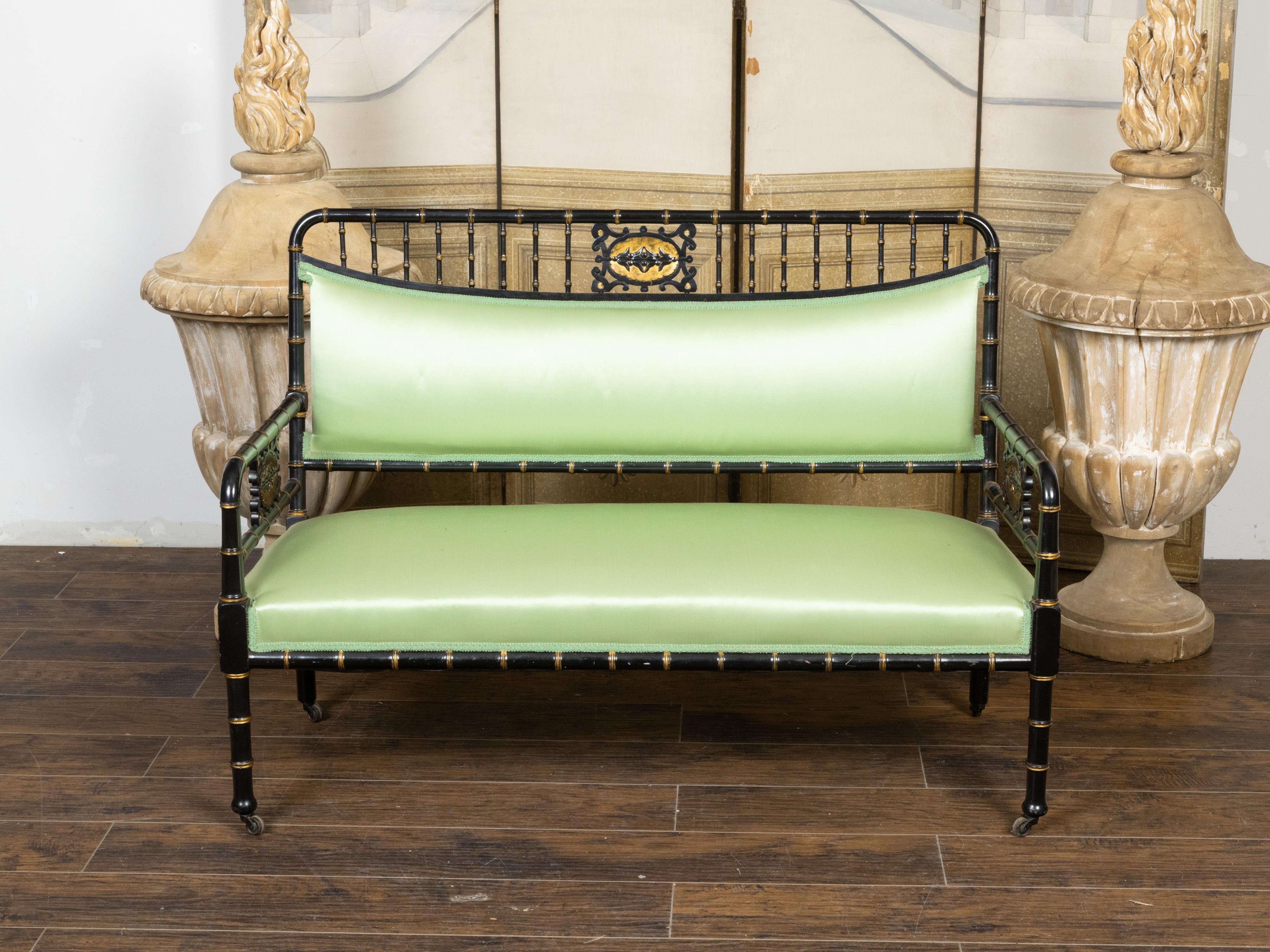 An English faux-bamboo bench on casters from the late 19th century, with ebonized finish, gilt accents, carved medallions and soft green upholstery. Created in England during the last quarter of the 19th century, this faux bamboo bench features a