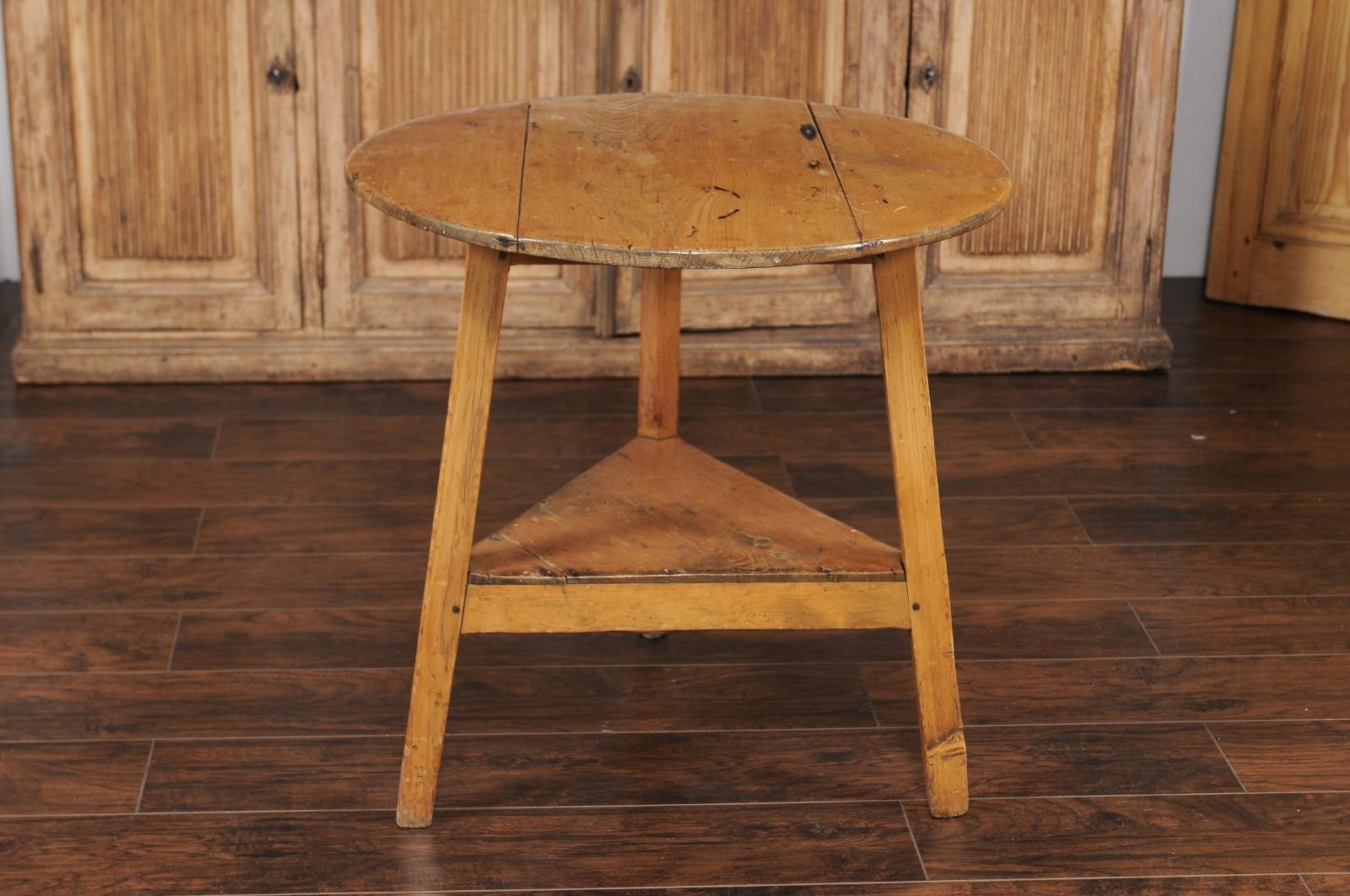 An English pine cricket table from the late 19th century, with circular top, blond finish and lower triangular shelf. Born in England in the last quarter of the 19th century, this cricket table features a circular planked top, resting upon a