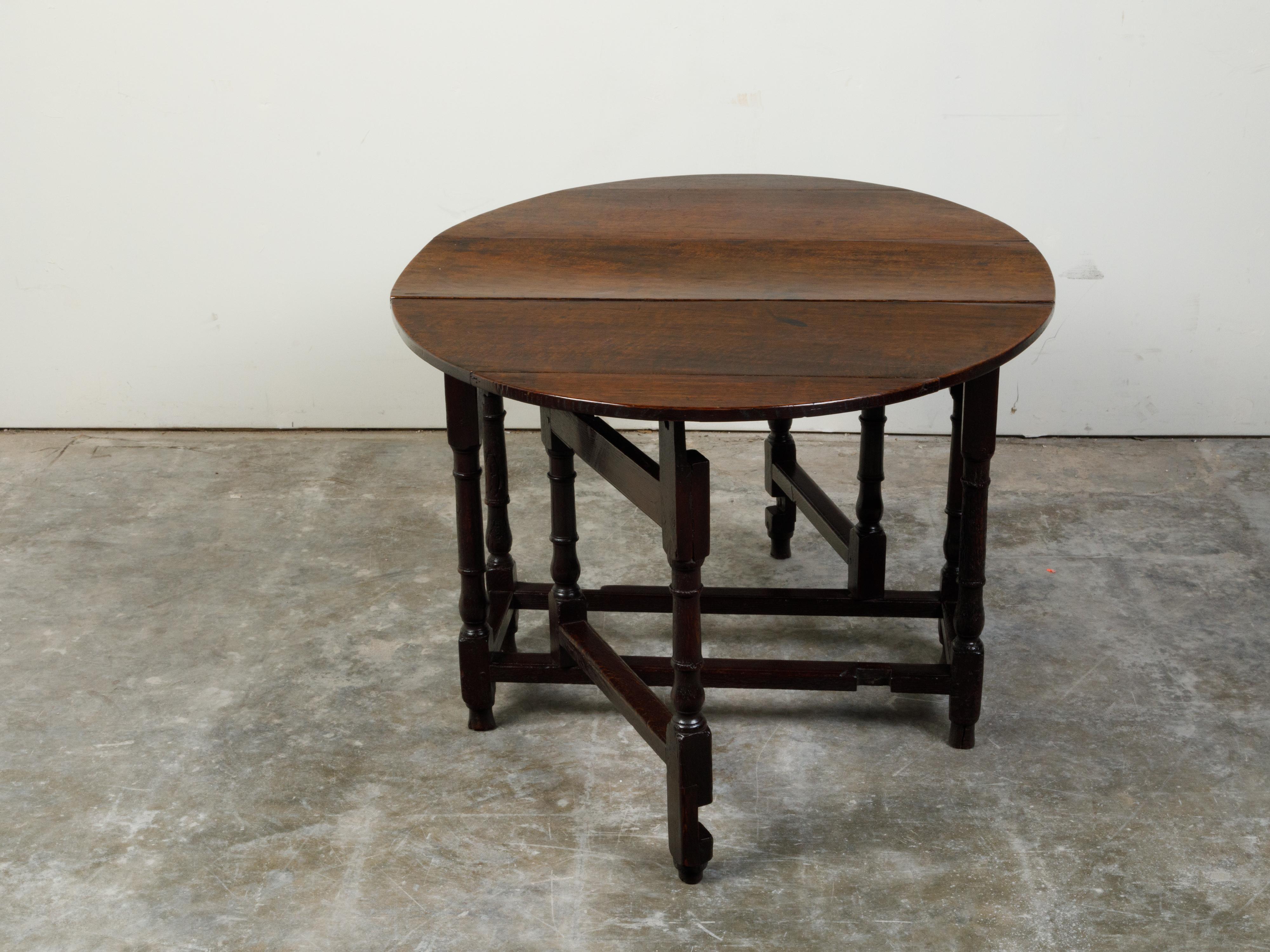 Une table rustique anglaise du 18ème siècle, avec un plateau ovale et des pieds tournés. Créée en Angleterre au XVIIIe siècle, cette table présente un plateau ovale composé de deux feuilles tombantes, reposant sur une base à pieds tournés. Accentuée