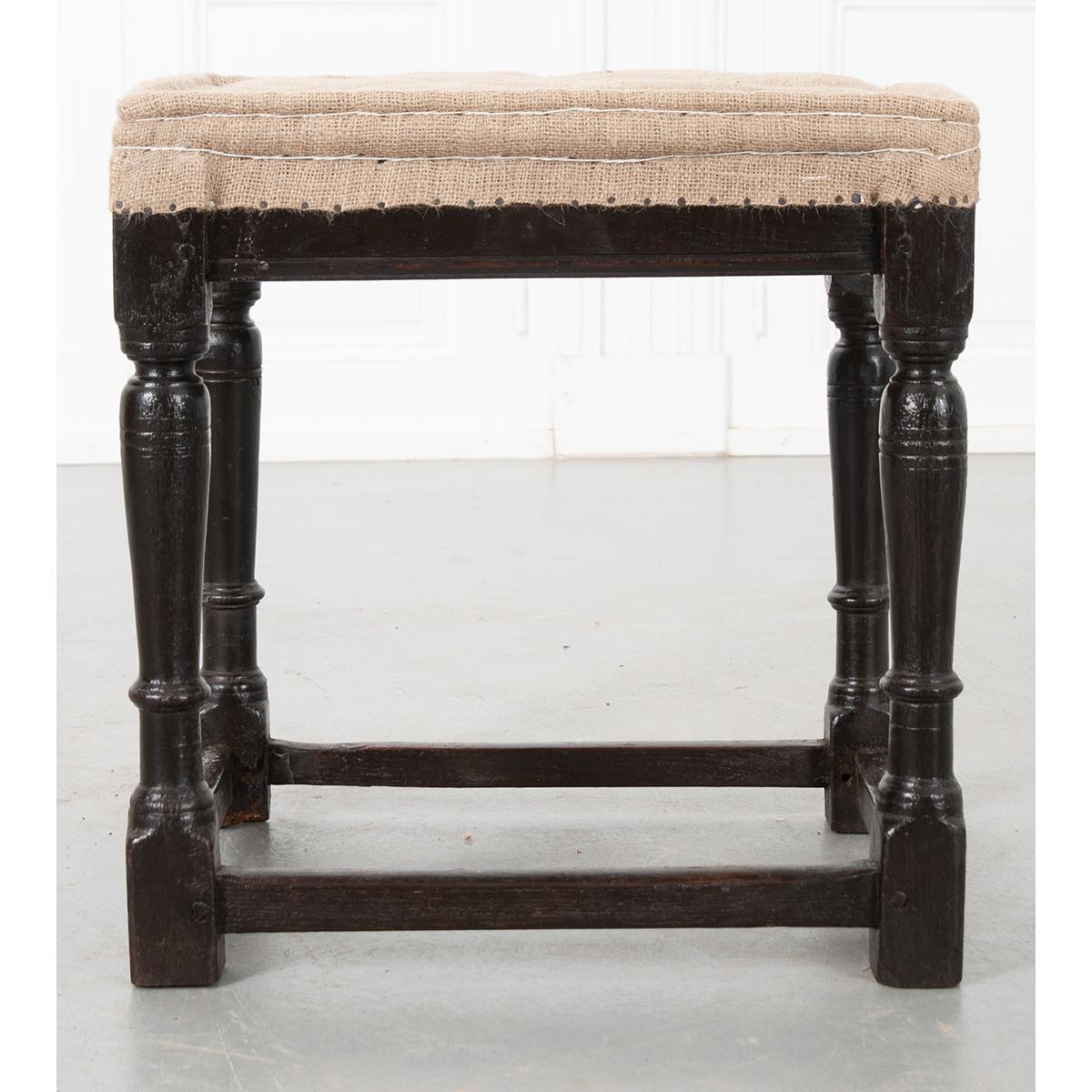 This is an English 18th century stool which has been painted black. It has burlap upholstery on the seat and can be used as it is or reupholstered. The four legs are turned and have stretchers around the bottom connecting all.
 