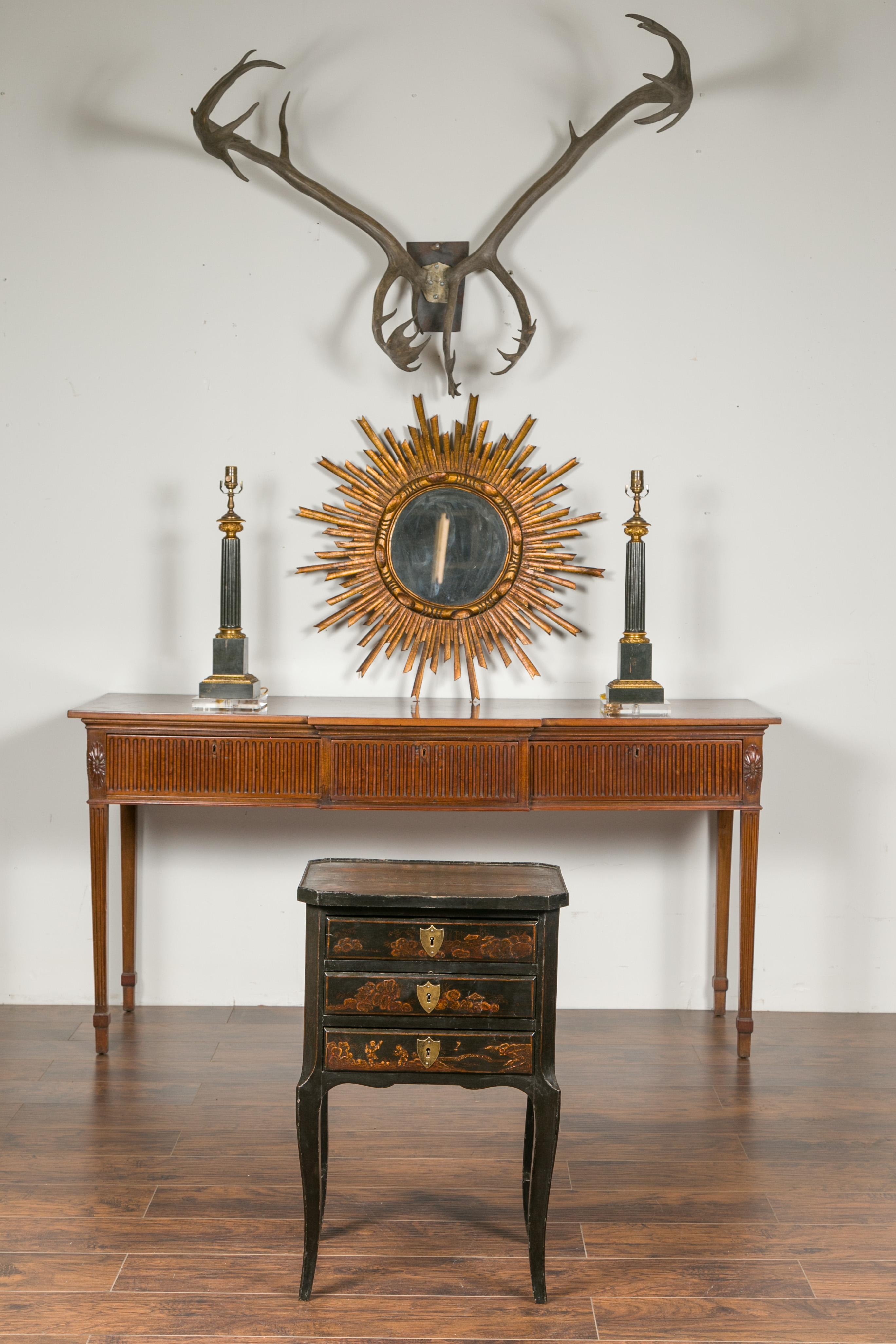An English black chinoiserie bedside table from the early 20th century, with three drawers and curving legs. Created in England at the turn of the century, this small bedside table feature a rectangular top with slightly raised edges, adorned with a