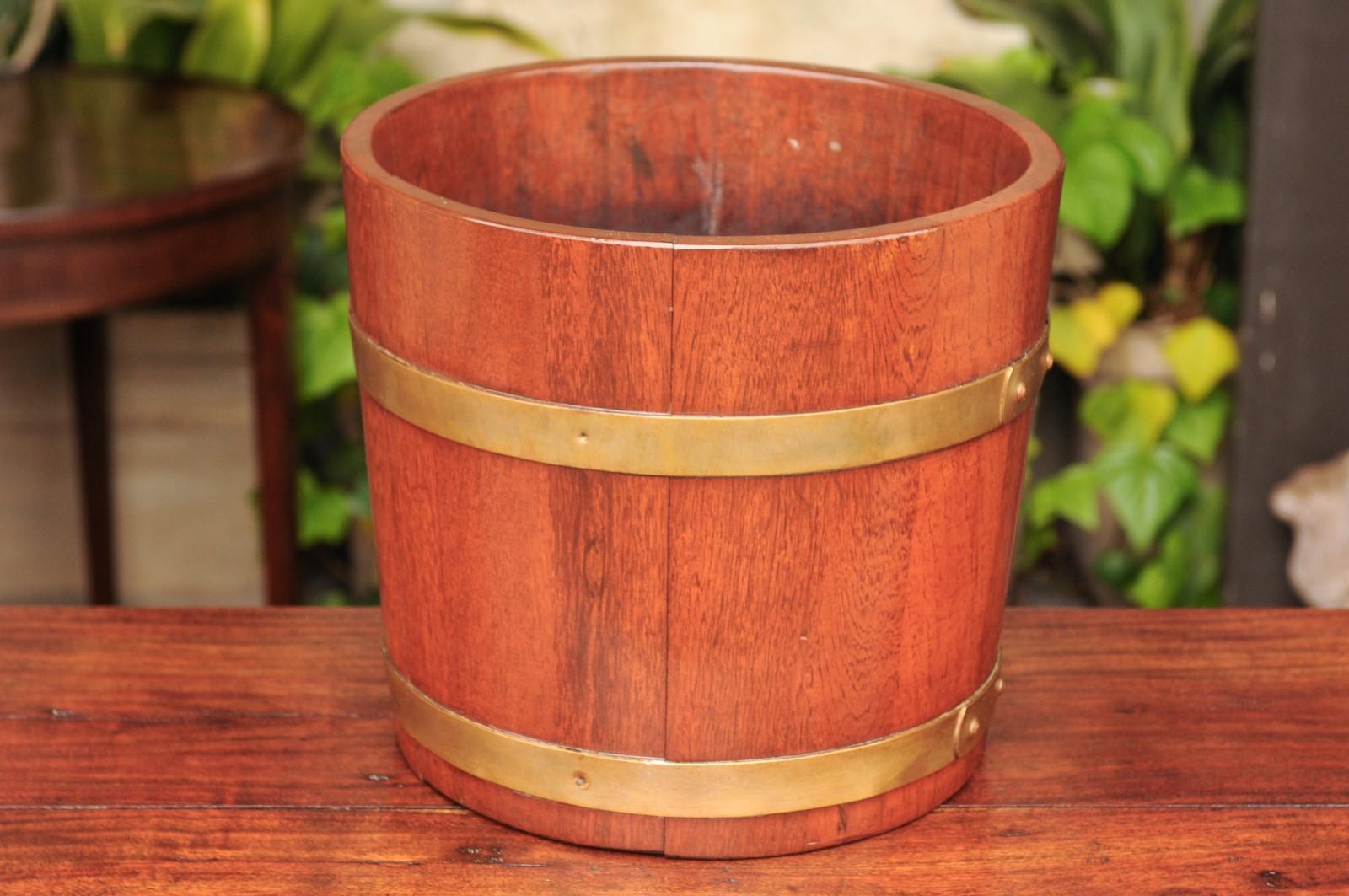 An English Edwardian period R.A Lister & Co. oak bucket from the early 20th century, with brass braces. Born in England in the early years of the short Edwardian reign, this charming bucket features a nice oak body, secured behind two horizontal