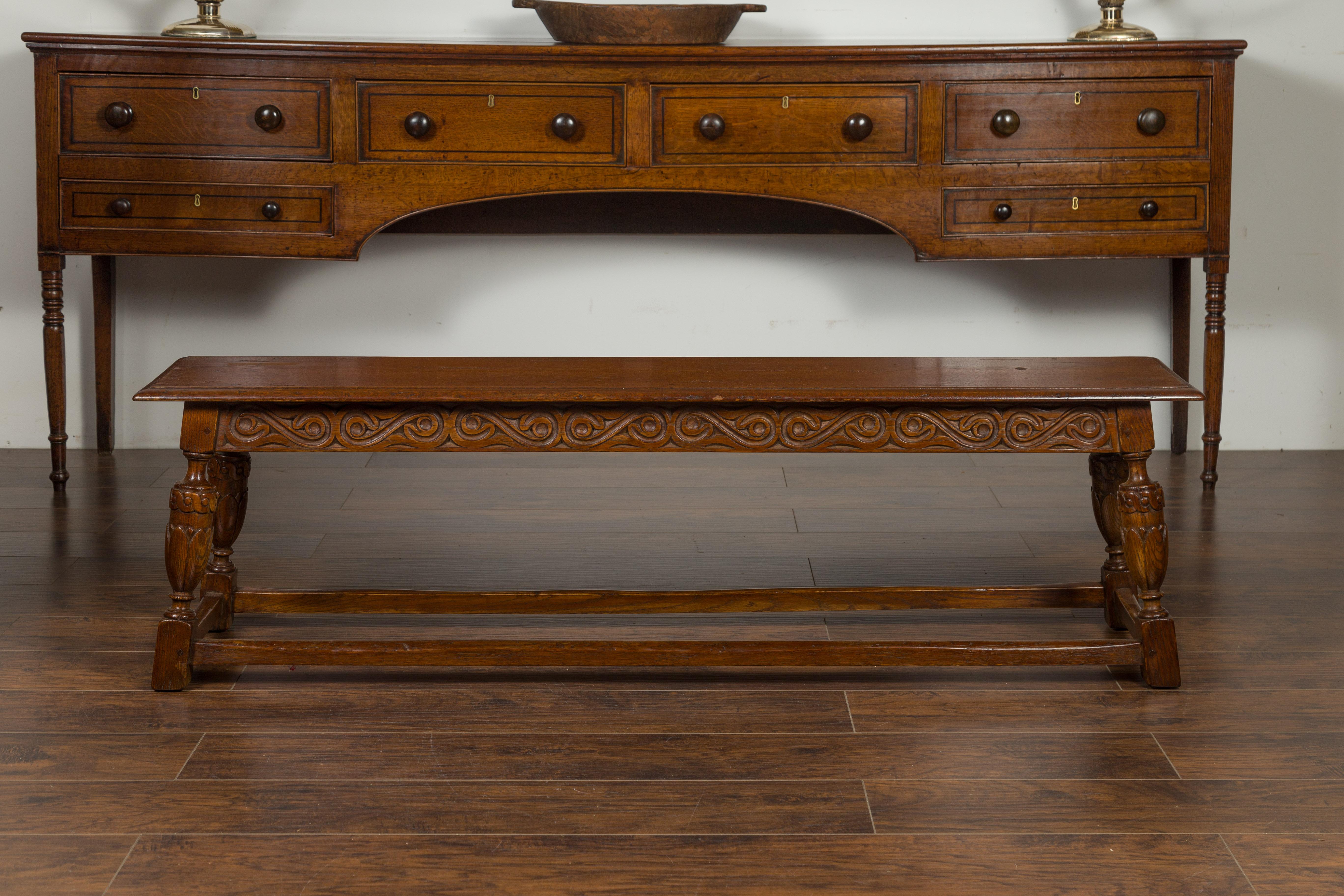 An English oak bench from the early 20th century, with carved apron and legs. Created in England at the turn of the century, this long oak bench features a rectangular top with beveled edges, sitting above an apron carved with scrolls on all sides.