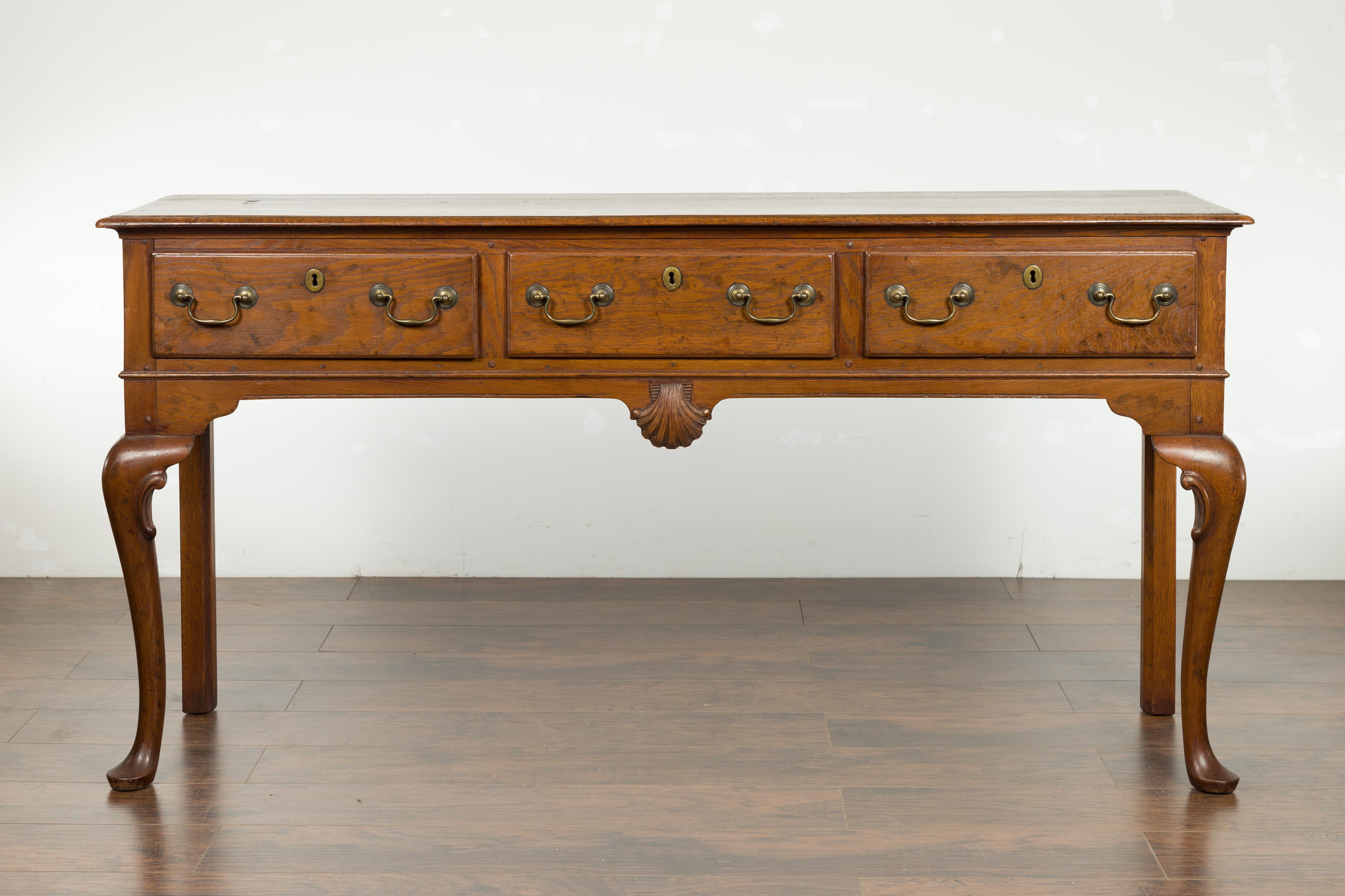 An English oak dresser base from the early 20th century, with three drawers, cabriole legs and carved shell motif. Created in England at the turn of the century, this oak dresser base / server features a rectangular planked top sitting above three