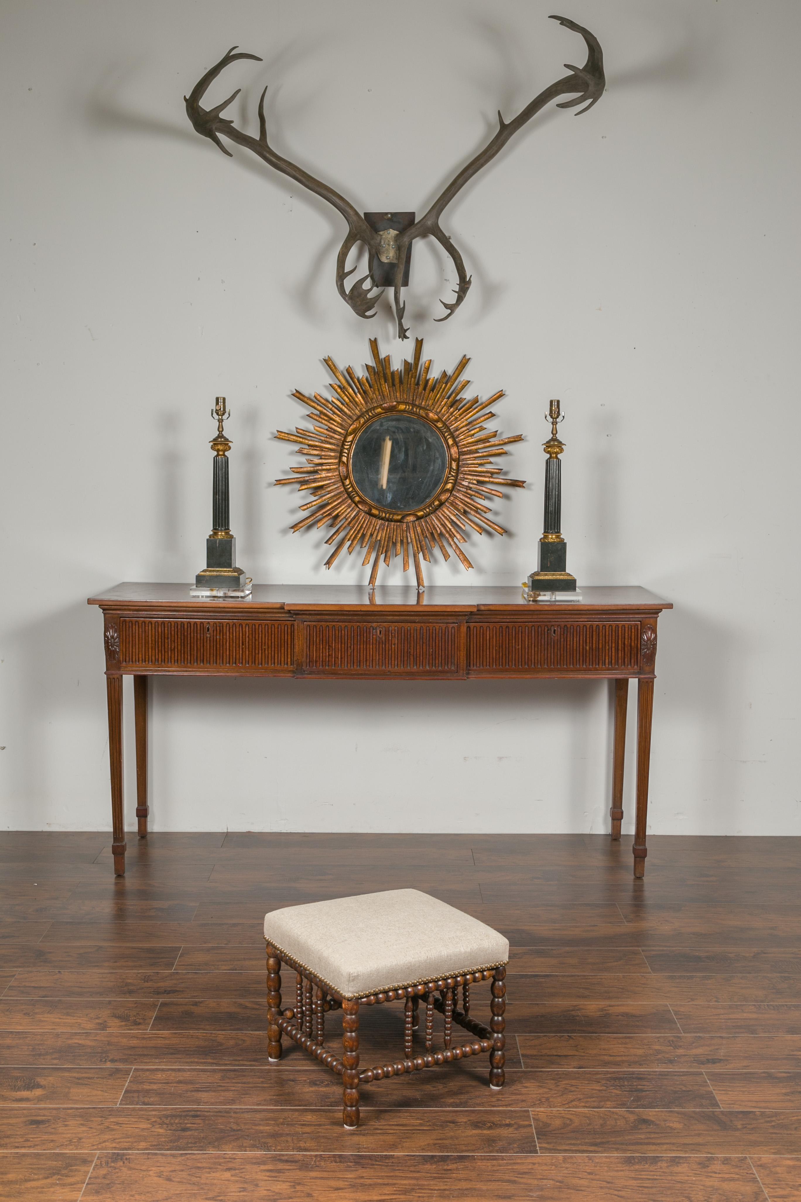 An English oak footstool from the early 20th century, with bobbin legs and new upholstery. Born in England during the turn of the century, this oak footstool features a square top covered with a linen fabric and nailhead trim, sitting above an
