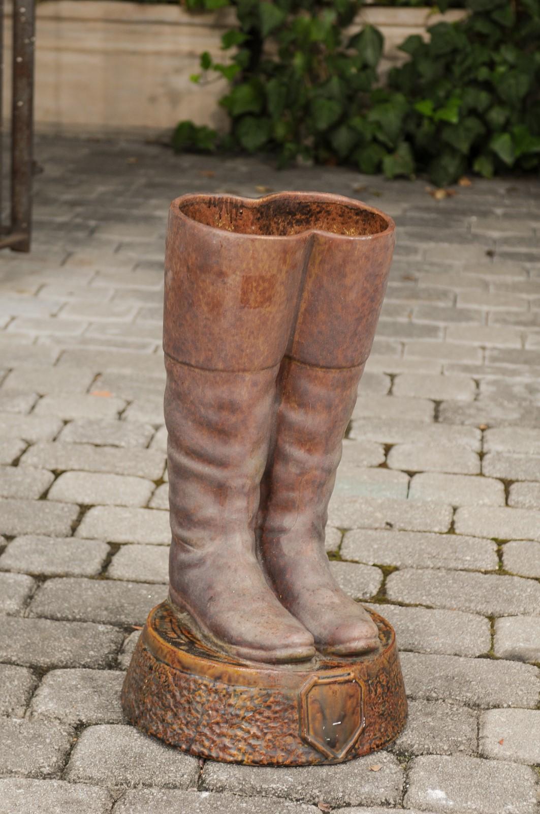  A contemporary terracotta umbrella stand from the early 21st century depicting boots resting on a circular base. This terracotta umbrella stand features a pair of brown riding boots resting on a circular tapering base, accented with a shield-shaped
