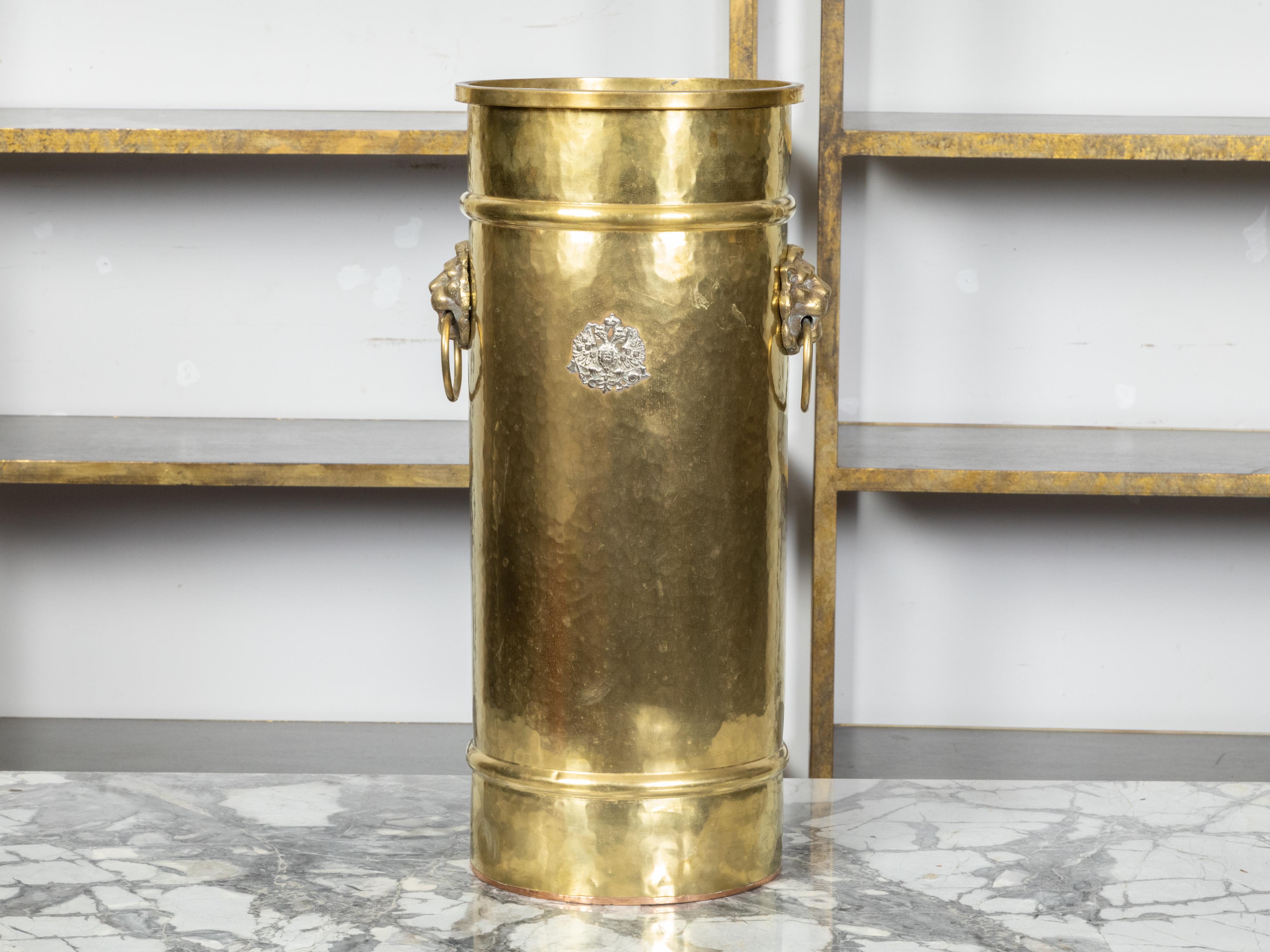 An English brass umbrella stand from the early 20th century, with lion ring pulls and heraldic coat of arms. Created in England during the Roaring Twenties, this umbrella stand features an oblong cylindrical body adorned with a double-headed eagle