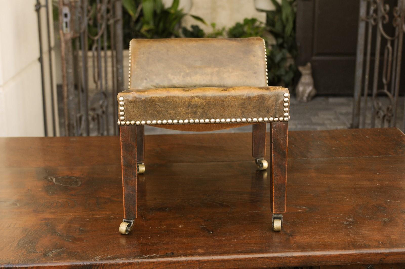 An English leather gout stool from the early 20th century, with out-scrolling back, nailhead trim and brass casters. Born in England during the second decade of the 20th century, this handsome gout stool features a brown leather upholstery with