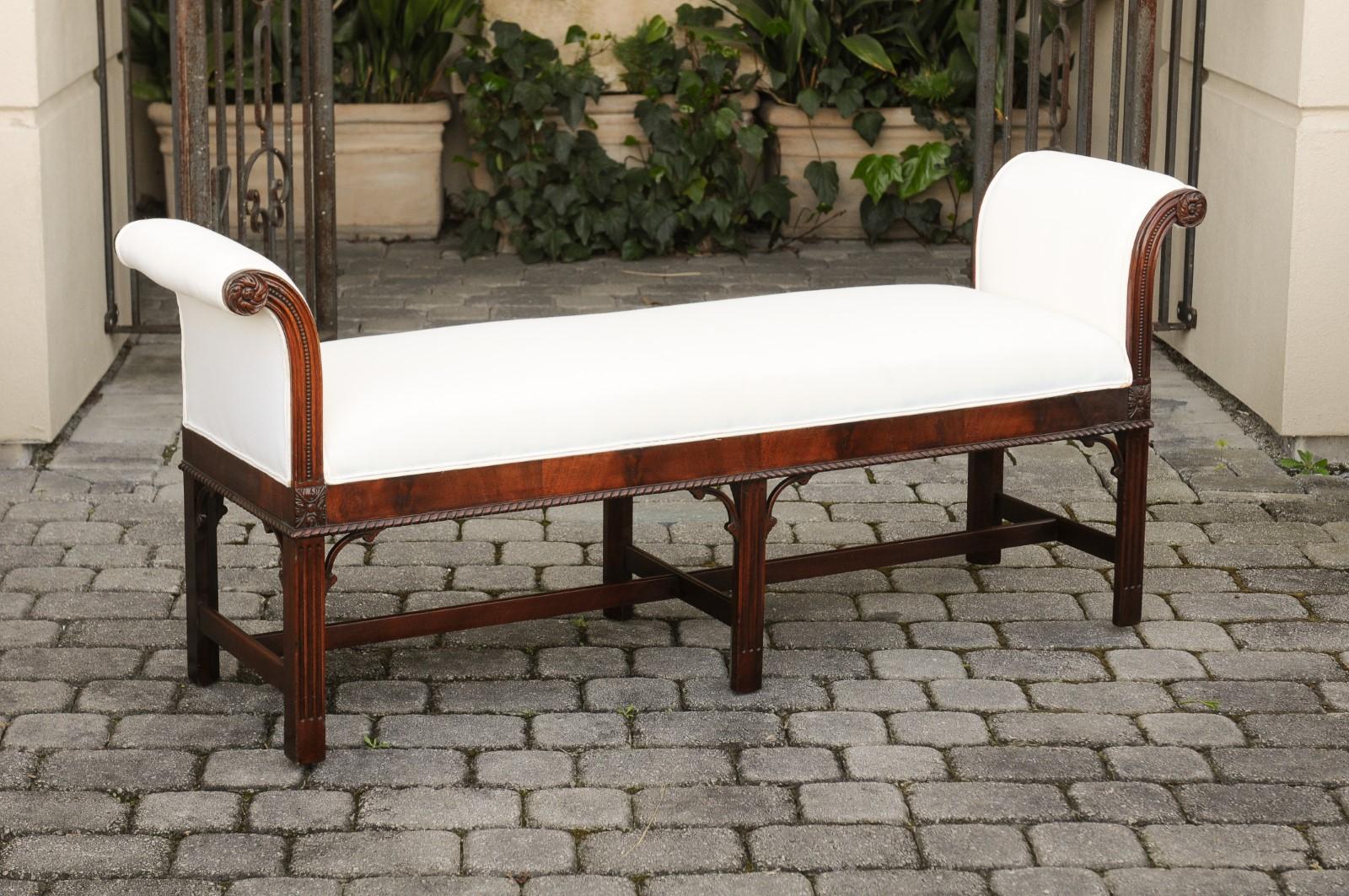 An English mahogany backless bench from the early 20th century, with out-scrolling arms, beaded and twisted motifs, carved rosettes and new upholstery. Born in England during the first half of the 20th century, this backless bench features a long