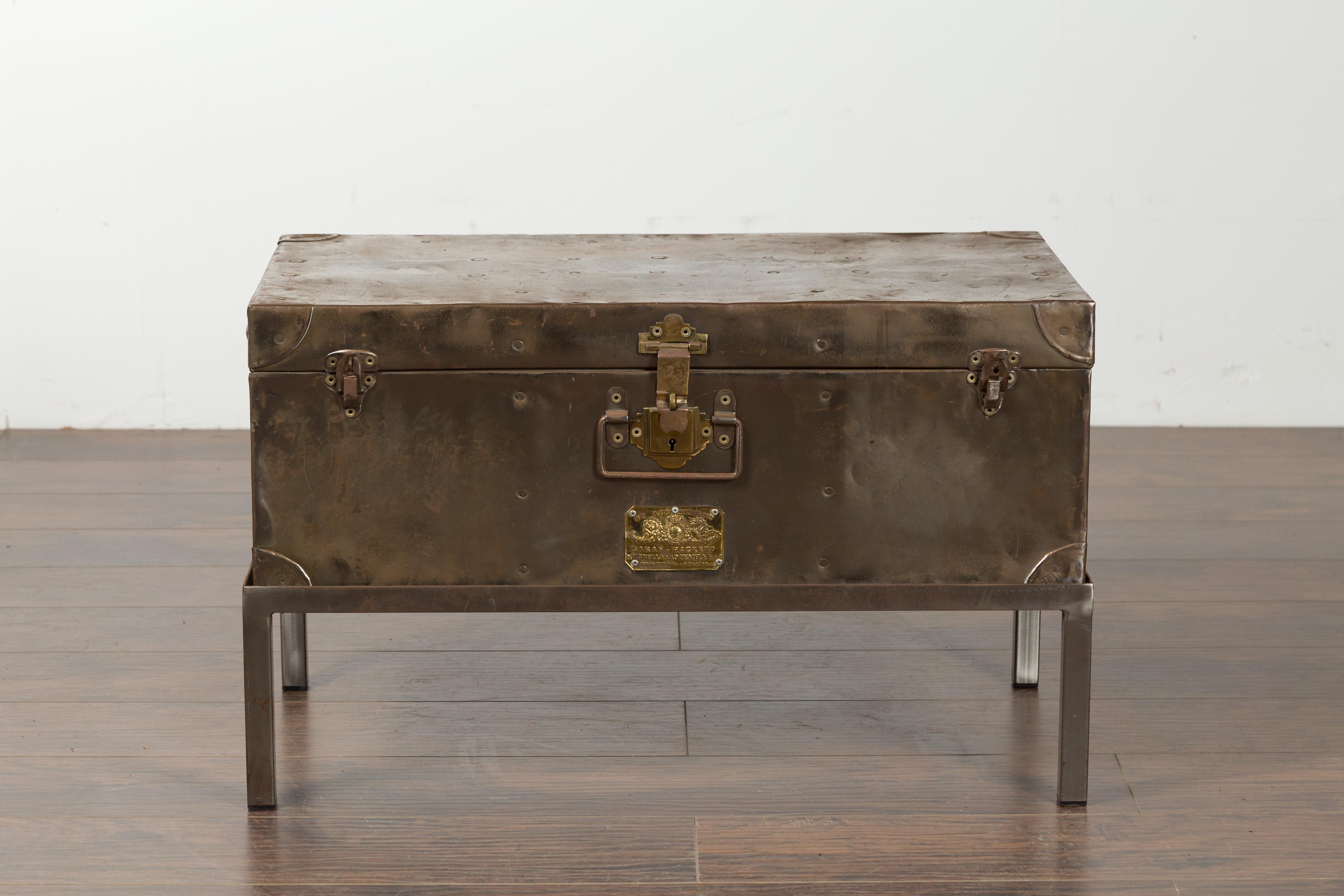 An English metal trunk from the early 20th century, on custom stand. Created in England during the first quarter of the 20th century, this metal trunk is accented with brass hardware. Marked by a brass plaque which says 
