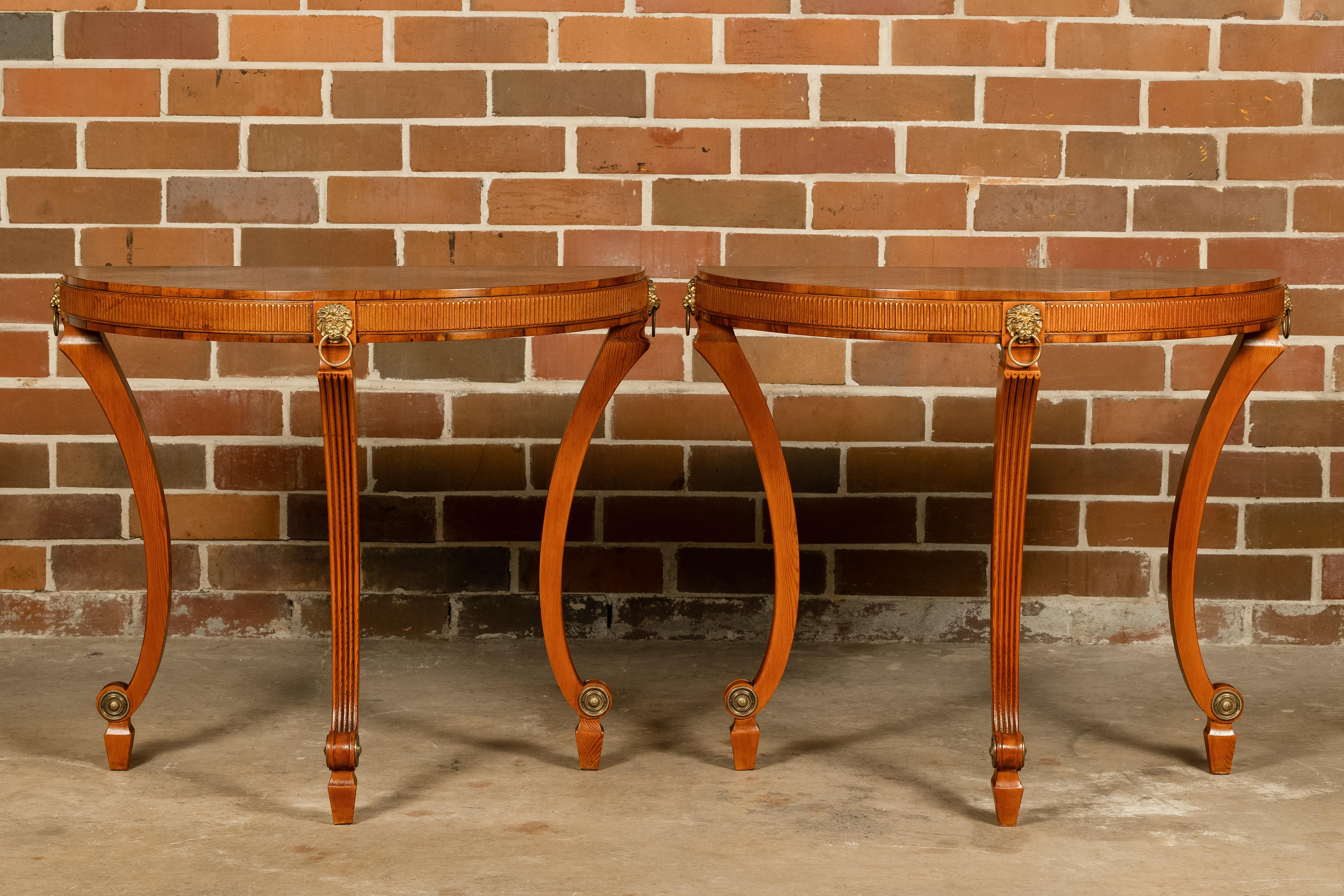 A pair of English Midcentury yew, pine and giltwood demi-lune console tables from circa 1960 with radiating veneered top, lion head ring pulls, fluted legs and apron, as well as scrolling feet. This pair of English Midcentury console tables, crafted