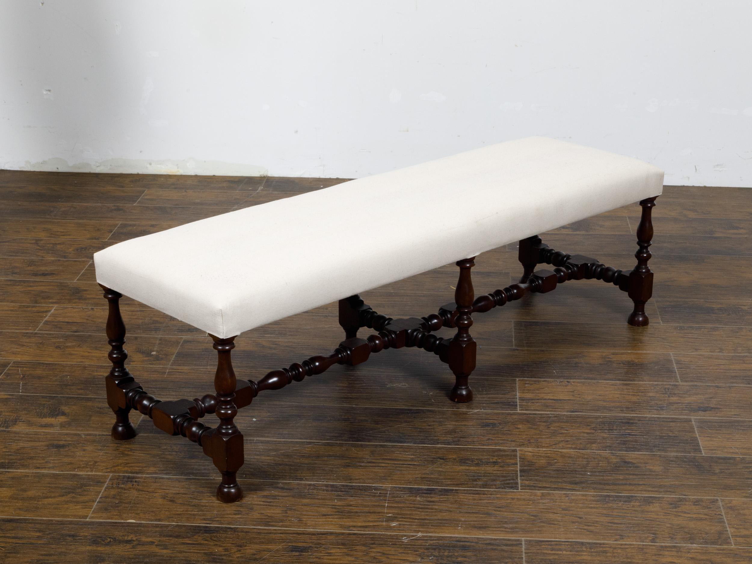 An English walnut bench from circa 1920 with six turned baluster legs, cross stretcher of similar style and new custom linen upholstery. This quintessentially English walnut bench from circa 1920 exudes timeless charm and sturdy elegance. The piece