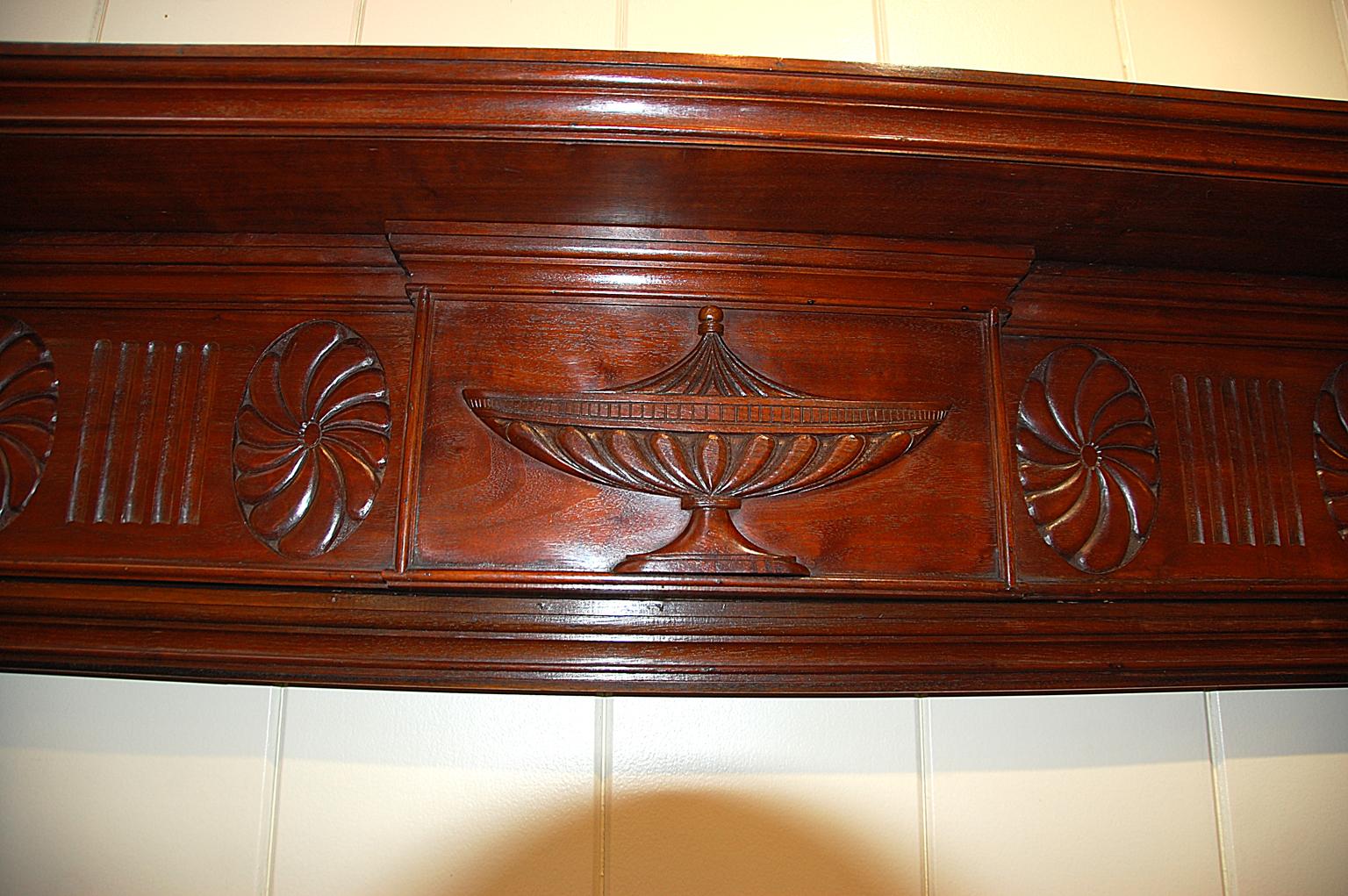 English Adam style 19th century carved mahogany fireplace surround and mantel. This elegant hand carved fireplace surround has a central urn motif with stylized flowers and anthemions framing it. The carved reeded columns surmounted by urns enhance