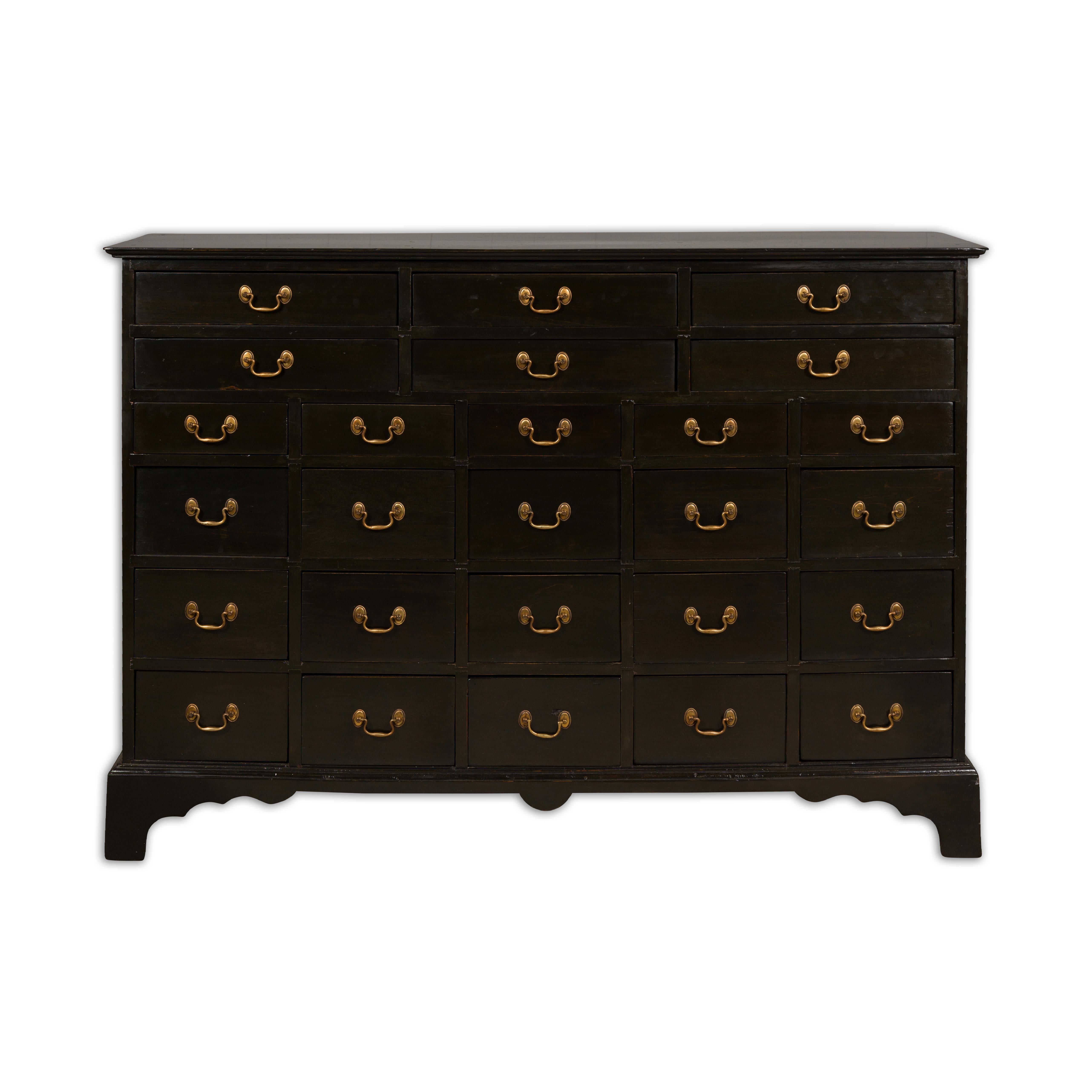 An English black apothecary chest from the 19th century with 26 drawers, brass hardware and carved bracket feet. Behold the grandeur of the 19th century with this English black apothecary chest, a piece rich in history and character. The chest,