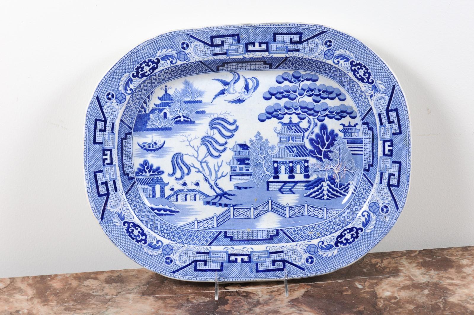 An English blue and white stoneware platter from the 19th century, with willow pattern and combed bottom. Born in England during the 19th century, this stoneware platter features a chinoiserie decor, presenting pagodas, trees, two birds and three