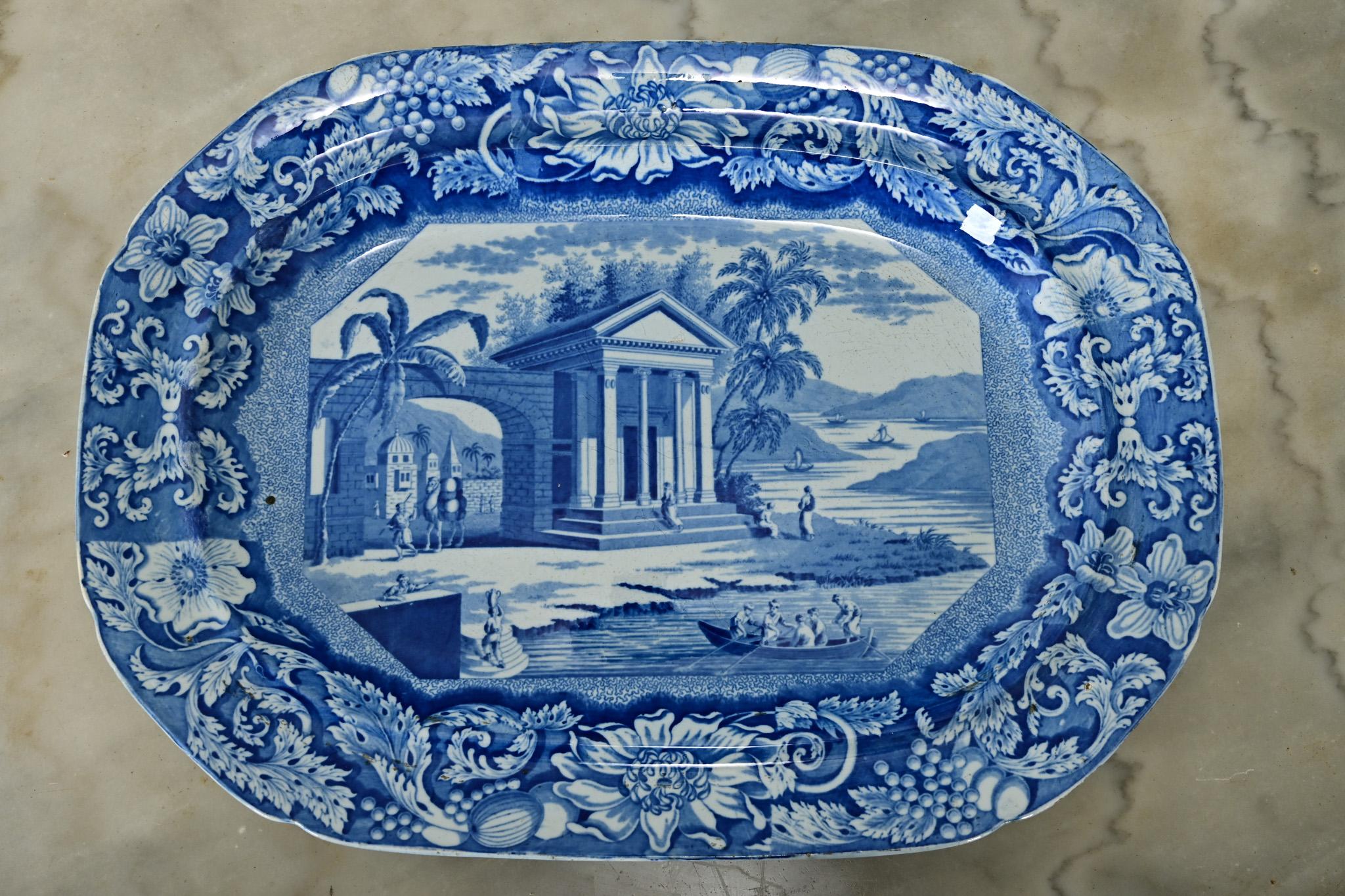 De taille généreuse, ce plat en céramique du XIXe siècle provenant d'Angleterre est décoré d'un motif bleu et blanc à la mode, conforme à l'époque. La forme de cartouche est encadrée par des fleurs exotiques le long du bord. L'image centrale
