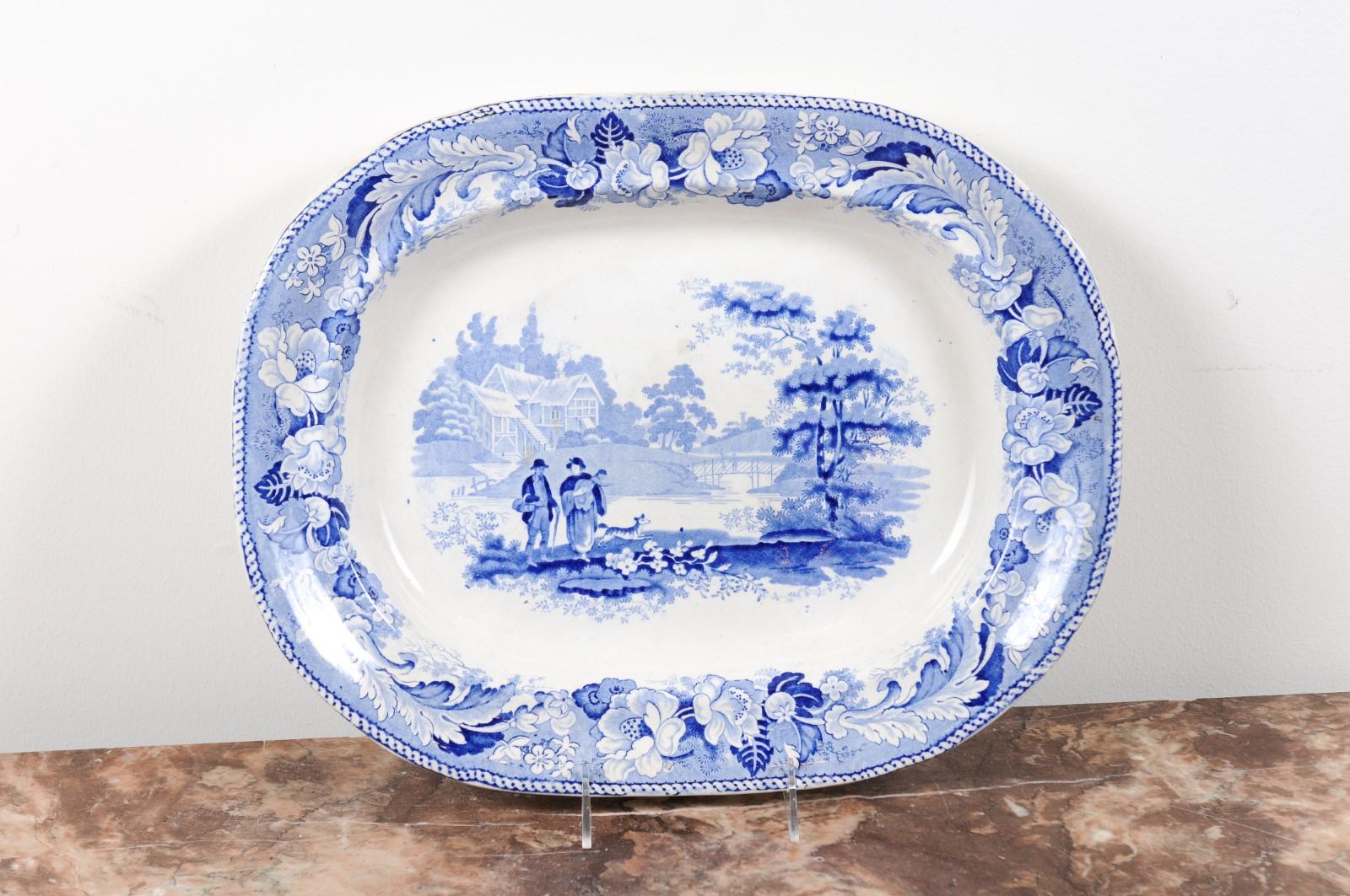 An English blue and white platter from the 19th century, with pastoral scene and floral border. Born in England during the 19th century, this blue and white platter features a peaceful pastoral scene. Two humble characters, accompanied by their dog,