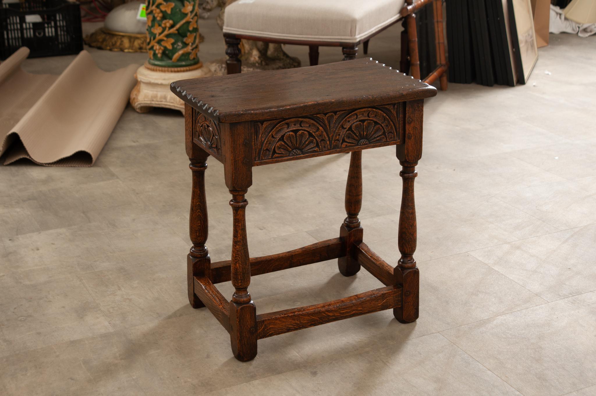 A lovely English oak joint stool with hand-carved motifs, 2 scalloped edges, and turned legs. Created in England in the 19th century, this oak joint stool features a rectangular top sitting above a hand-carved apron. The ensemble is resting on four