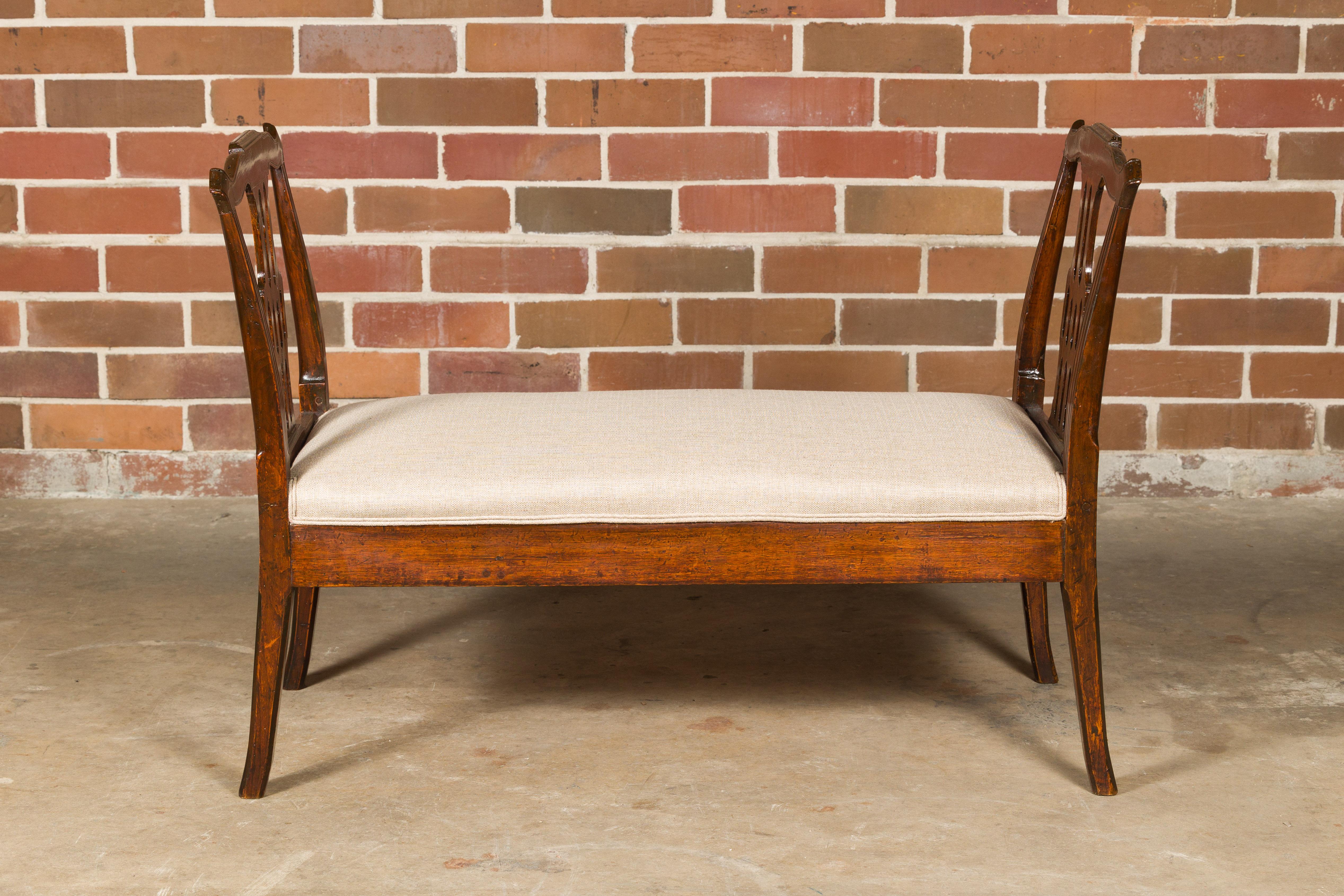 An English wooden bench from the 19th century with carved side supports, saber legs and new custom linen upholstery. This 19th-century English wooden bench is a timeless blend of elegance and functionality. Its classic design, with gracefully carved
