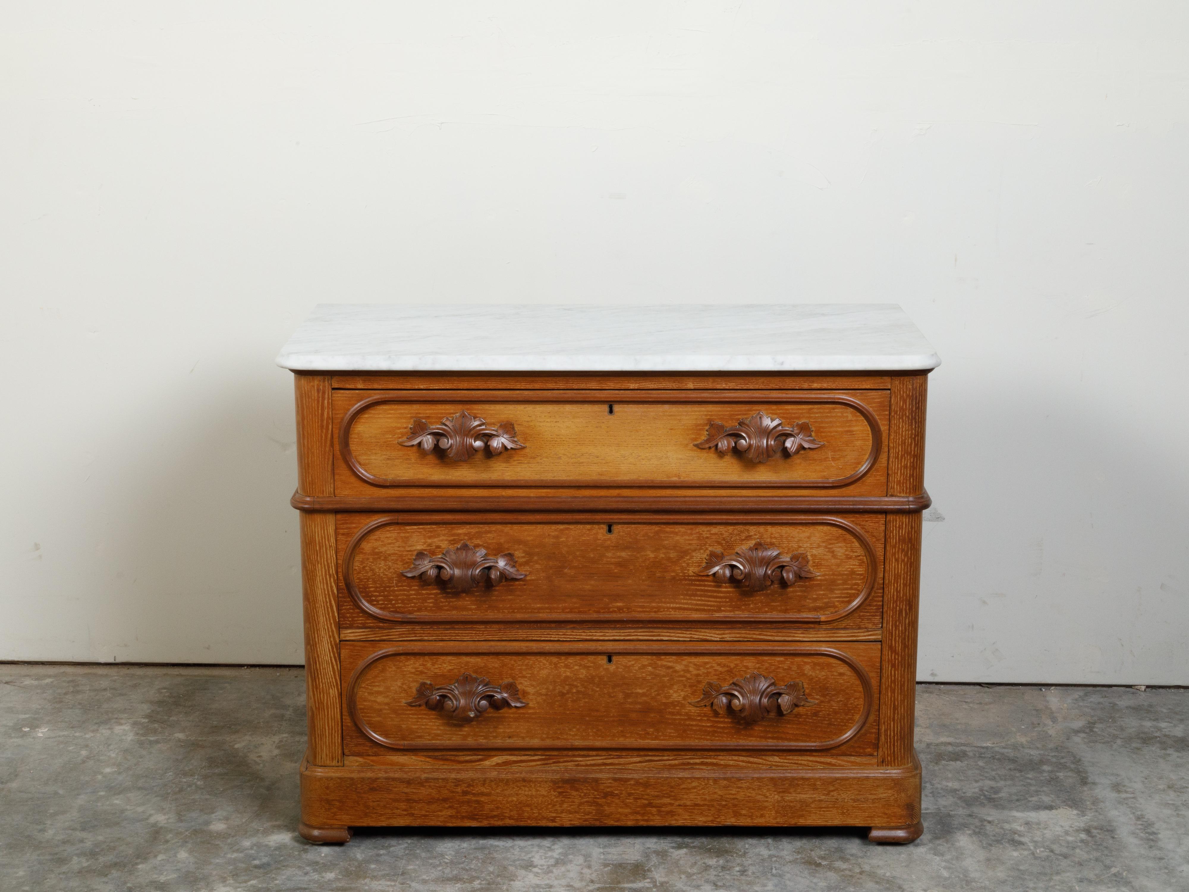 An English chestnut three-drawer chest from the late 19th century, with white marble top and foliage-carved handles. Created in England during the last quarter of the 19th century, this chest features a rectangular white marble top with rounded