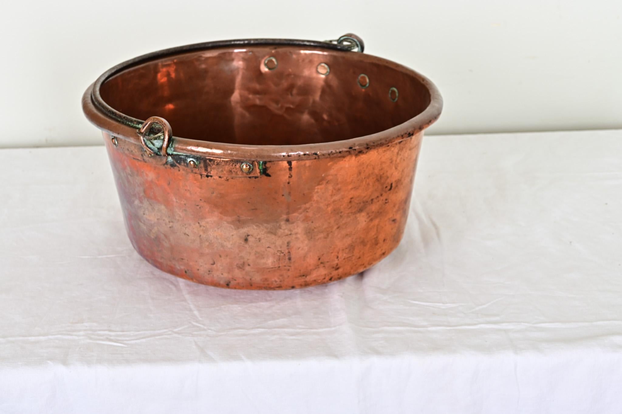 Une ancienne marmite anglaise en cuivre, avec une poignée en fer pour la suspendre au-dessus du feu, datant d'environ 1850 ou plus tôt. L'extérieur présente une belle patine, assombrissant la texture martelée et accentuée par des fleurs de