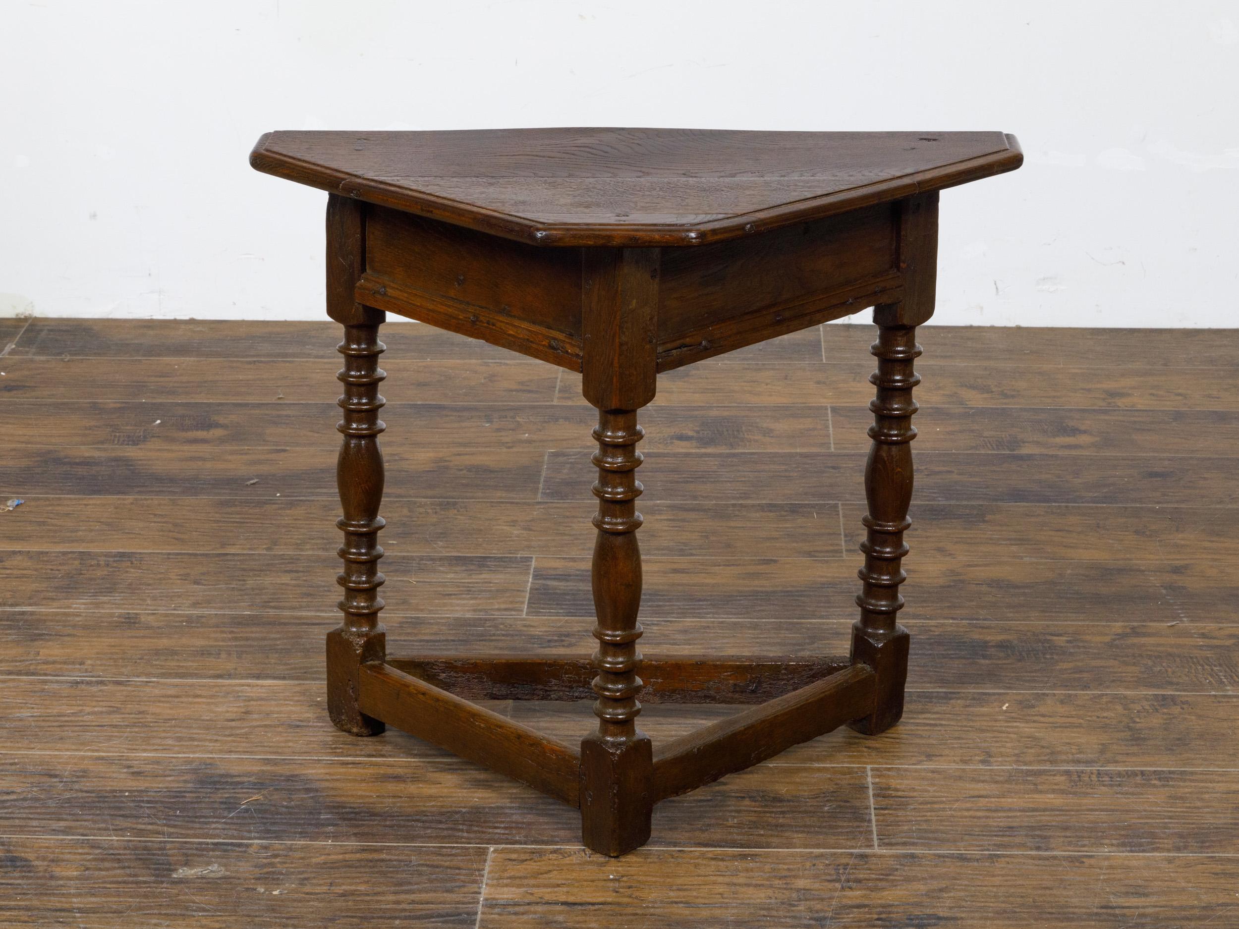 An English triangular shaped oak demi-lune table from the 19th century with turned legs and plain side stretchers. This English demi-lune table, crafted from dark oak, features a triangular-shaped top with a flat frontal section, supported by