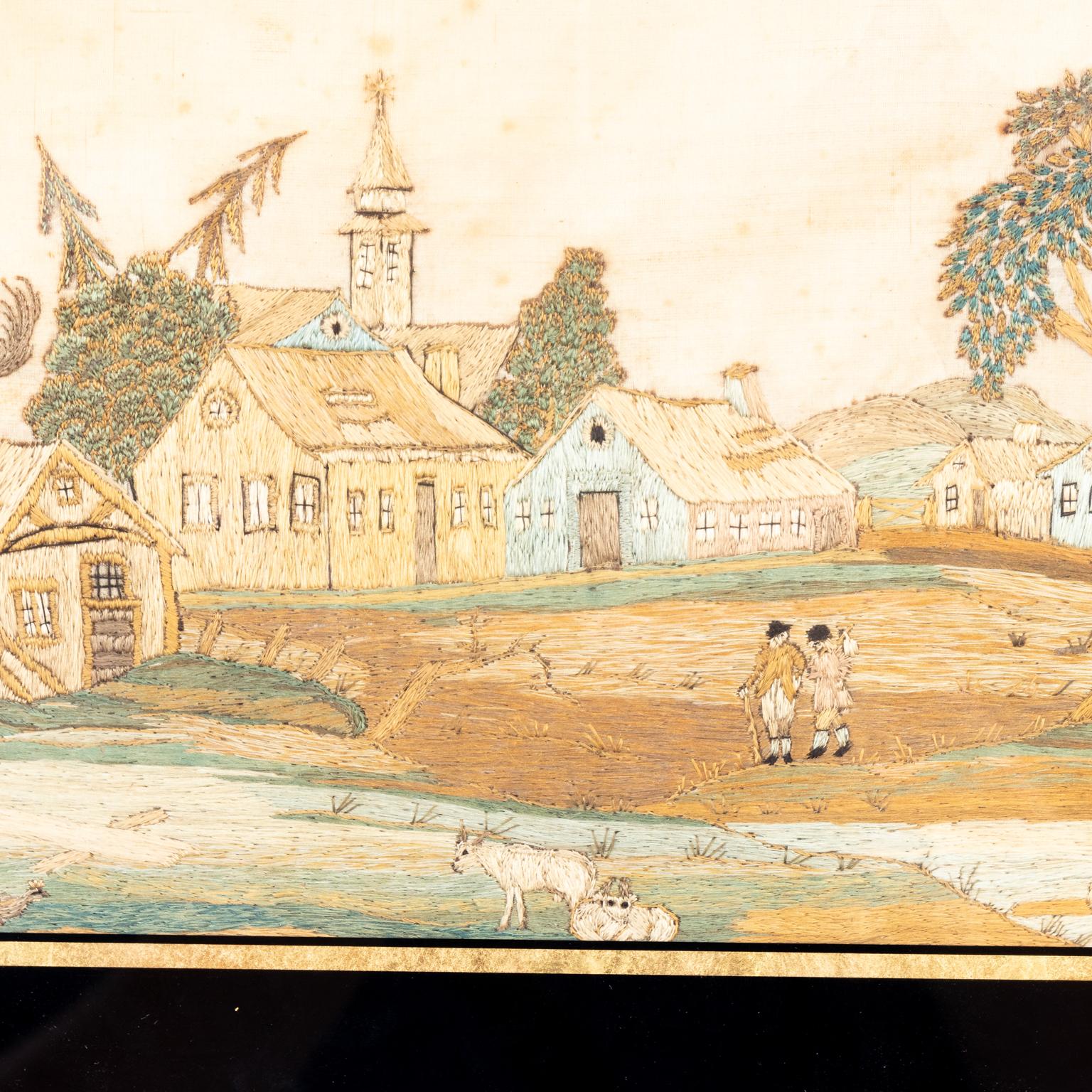 Circa 1825 Georgian style silk embroidered country scene in gilt frame detailed with two figures and two sheep in the foreground framed by rustic buildings and a steepled church in the background. Made in England. Please note of wear consistent with